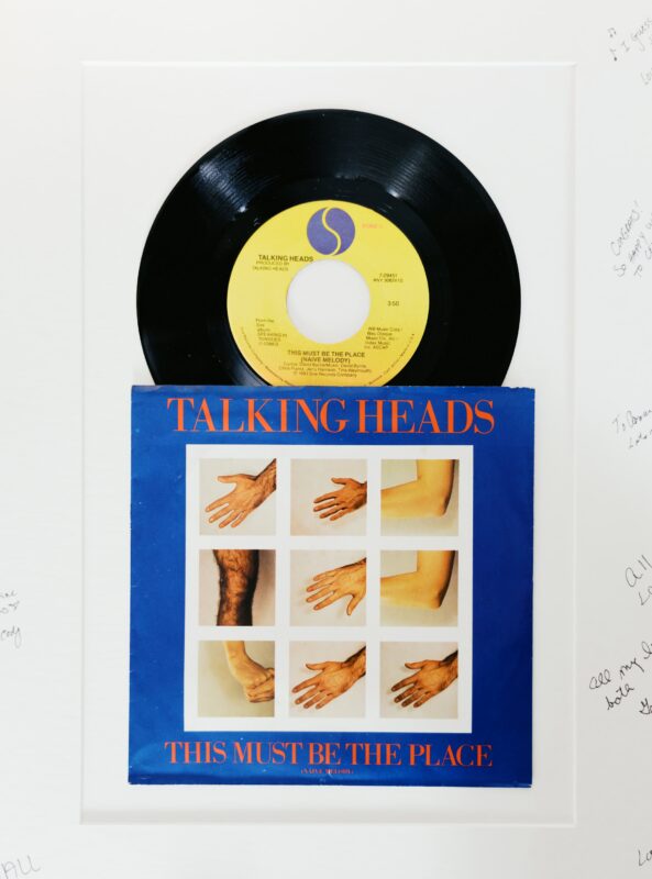 A vinyl record and its sleeve for Talking Heads' "This Must Be the Place (Naive Melody)" are displayed on a white mat with signatures for a unique wedding guestbook. The sleeve shows images of various hands.