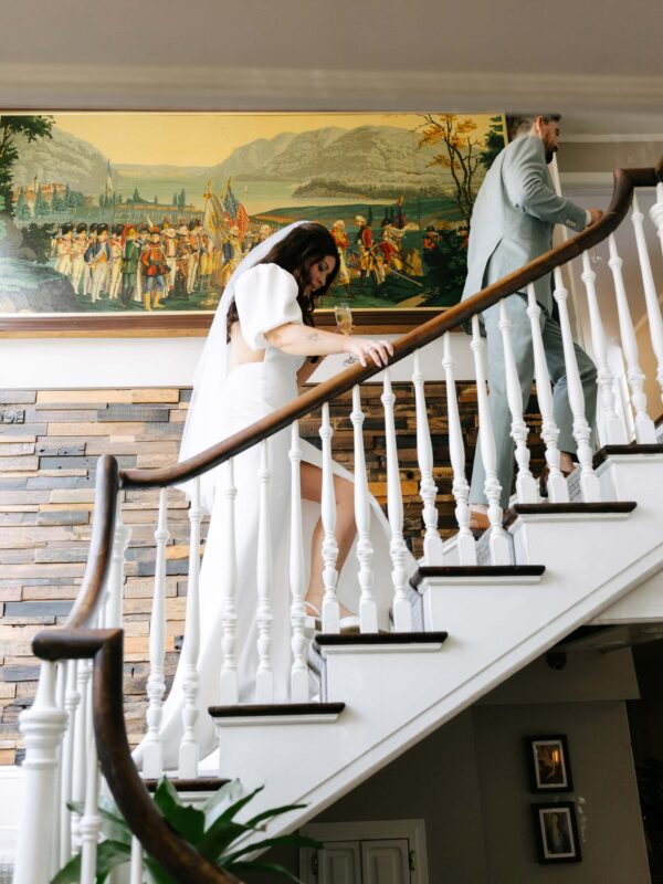 A bride in a white dress and veil walks up a spiral staircase behind a man in a suit, with a historical painting on the wall above them.