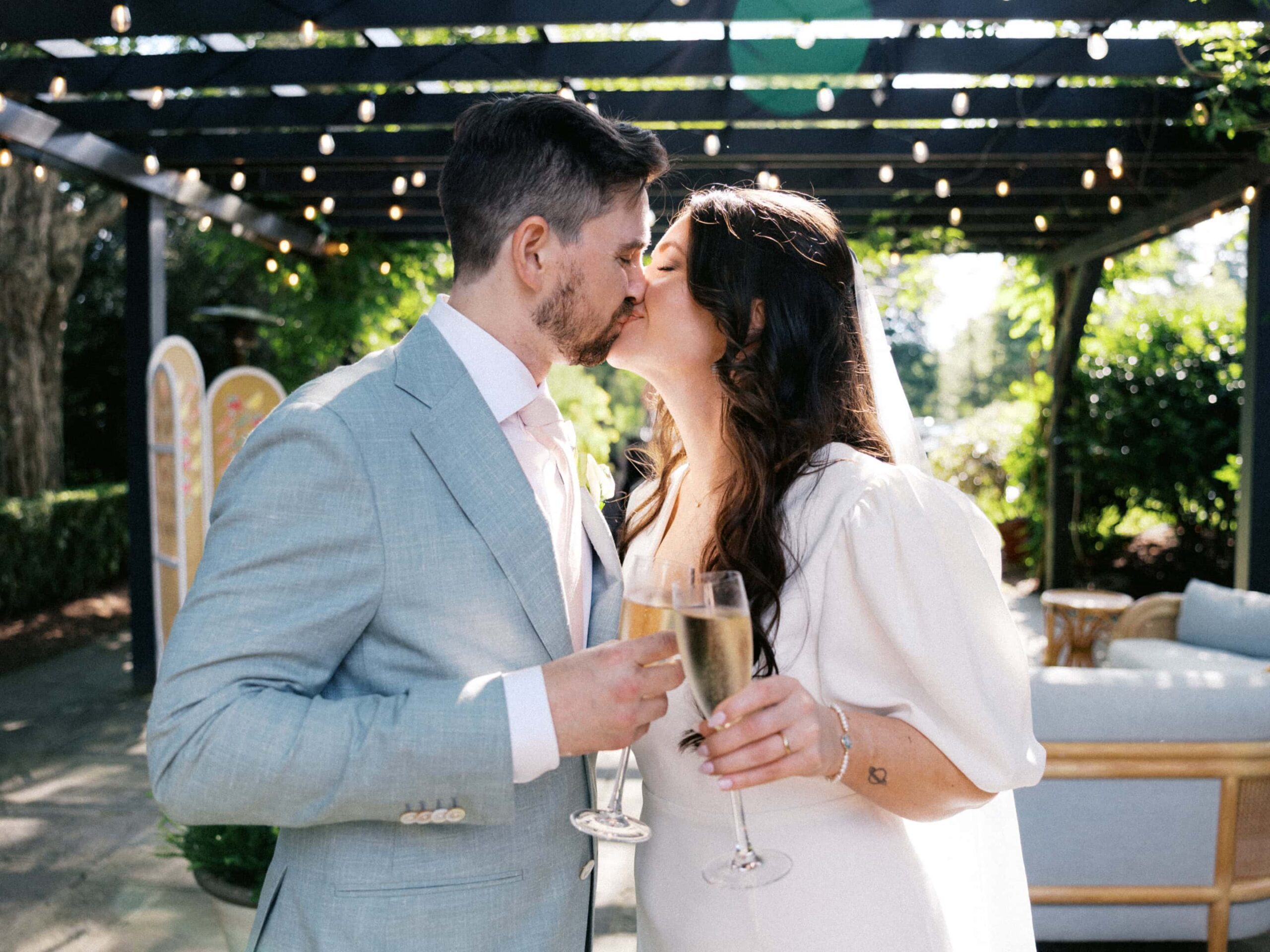 A bride and groom kiss under string lights with champagne during an outdoor cocktail hour.
