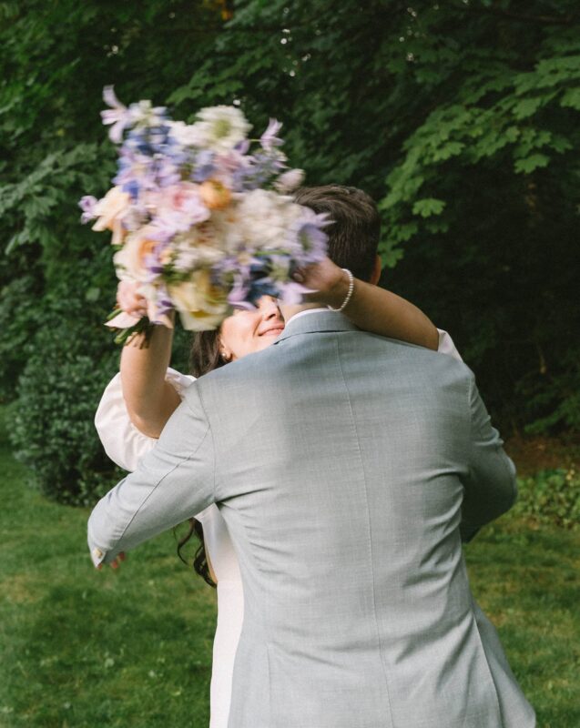 A couple embraces outdoors; the woman's face is partially hidden by a blurry bouquet of flowers she holds in front of them.