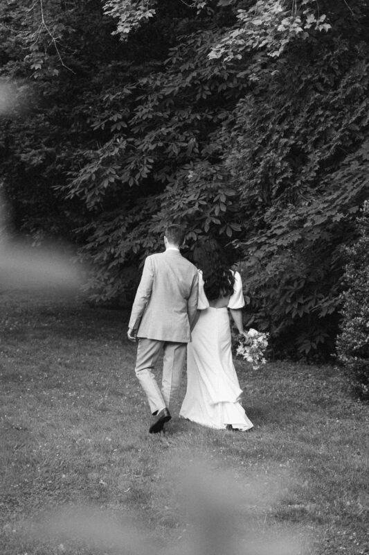 A married couple, dressed in formal attire, walks hand in hand on grass toward dense trees. The image is in black and white.