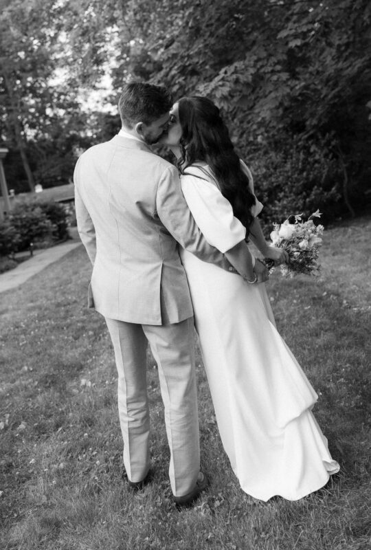 A couple dressed in formal attire stands on grass, embracing and kissing, with the woman holding a bouquet behind her back. The scene is outdoors and photographed in black and white.