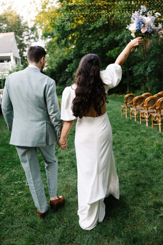 A bride and groom walk hand in hand on grass; the bride holds a bouquet aloft. Empty chairs are arranged in rows outdoors, string lights are overhead.