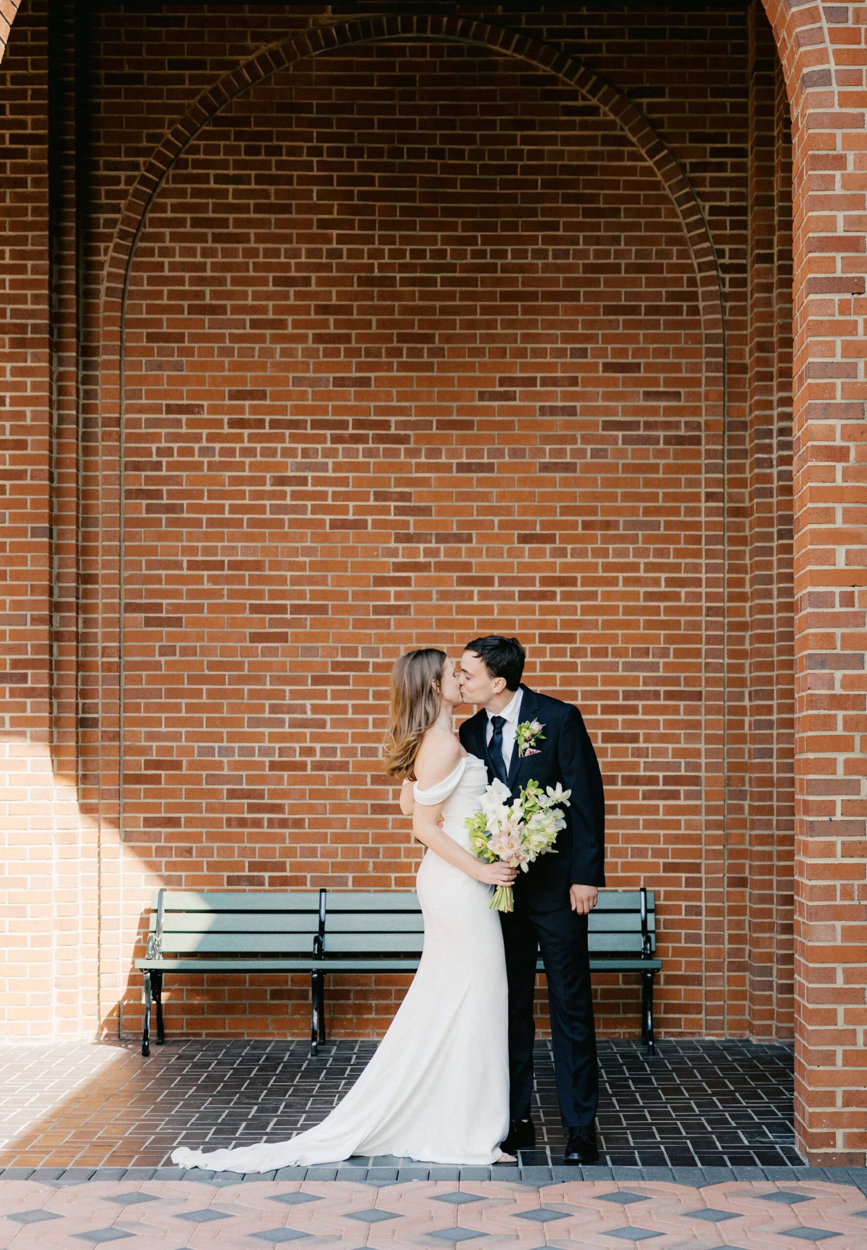 A bride and groom kiss in an arched brick portico.