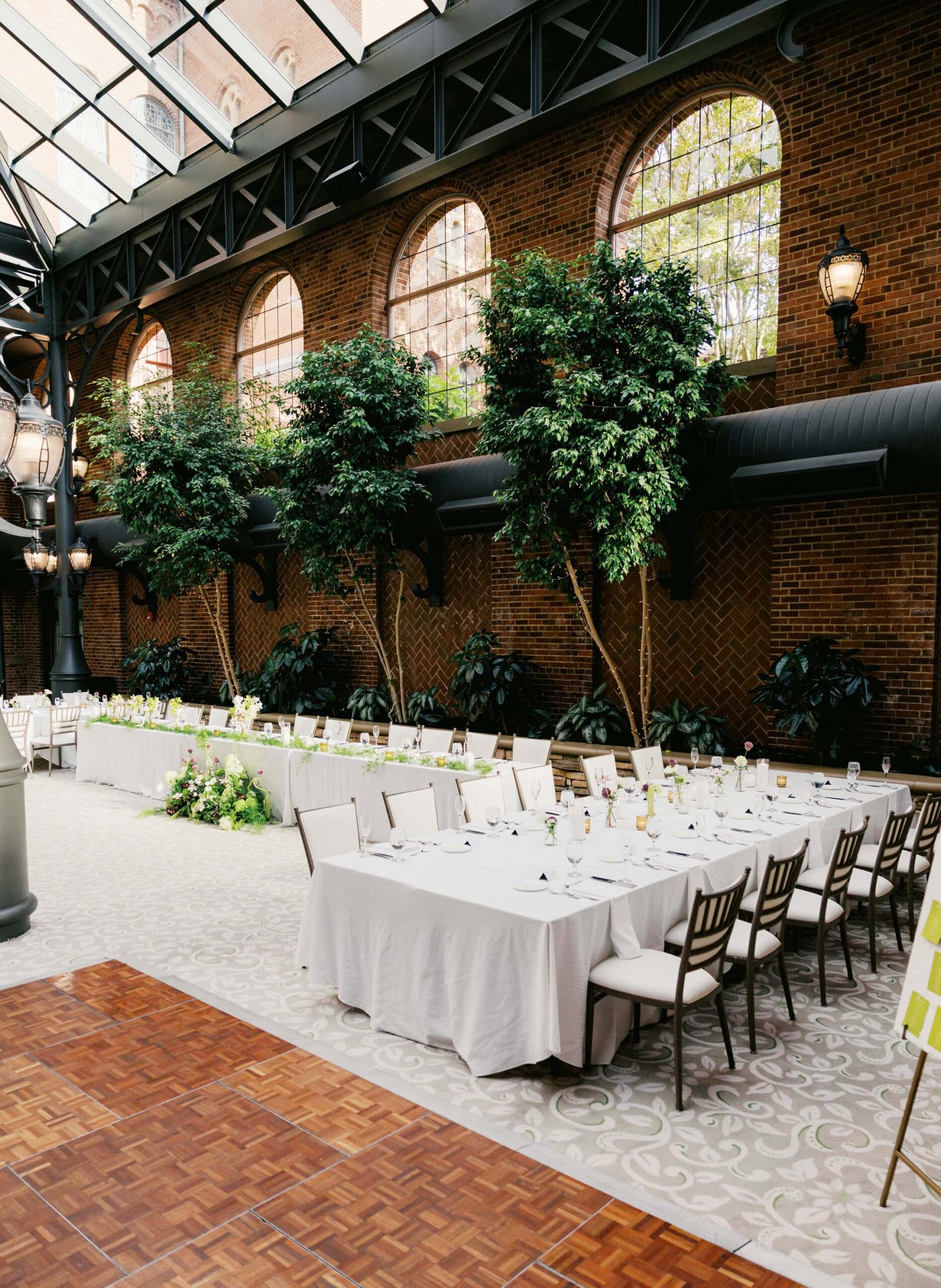 An open air atrium wedding venue with a glass ceiling, arched windows, and indoor trees lining red brick walls.