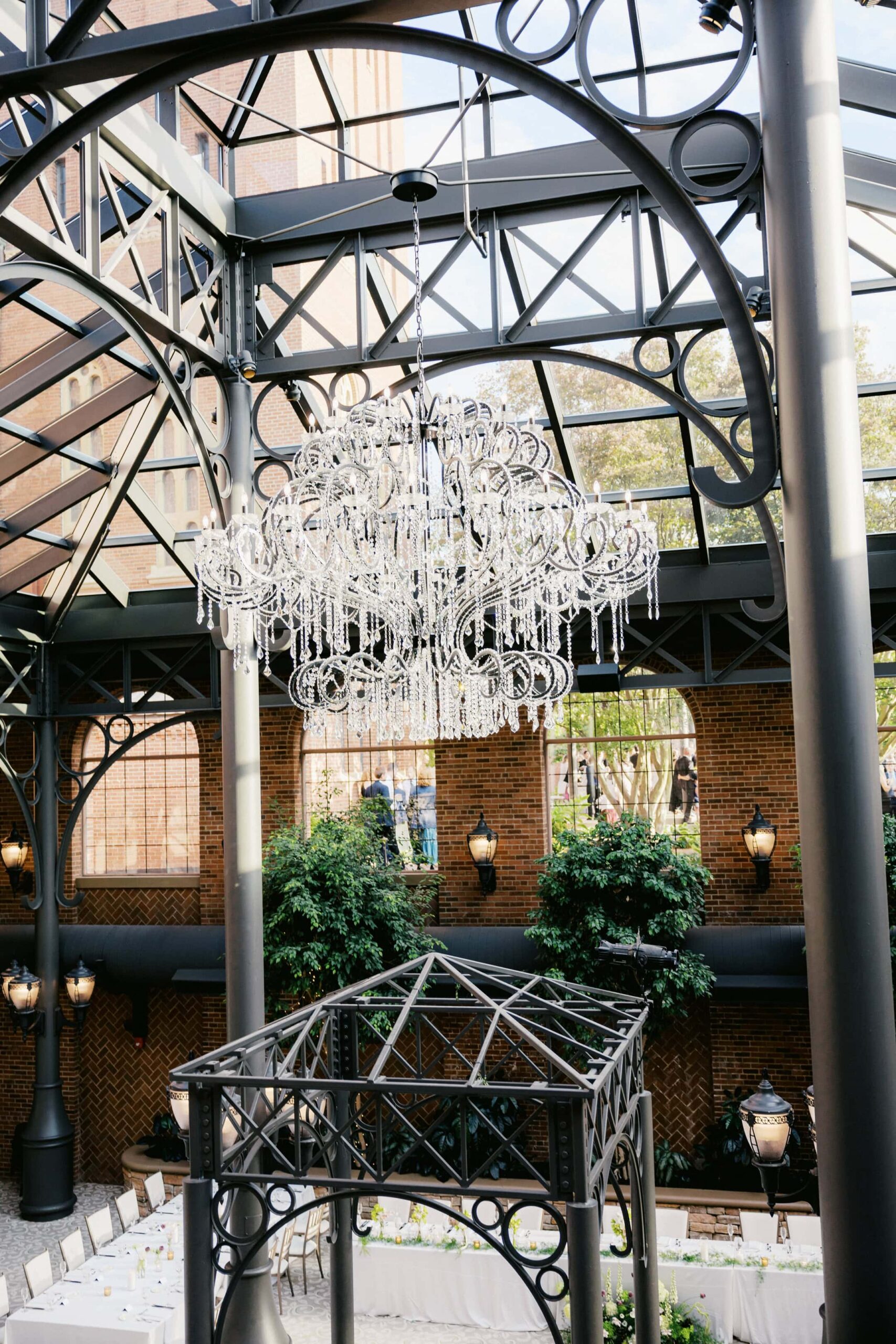 An open air atrium wedding venue with a glass ceiling, crystal chandelier, and indoor trees lining red brick walls.