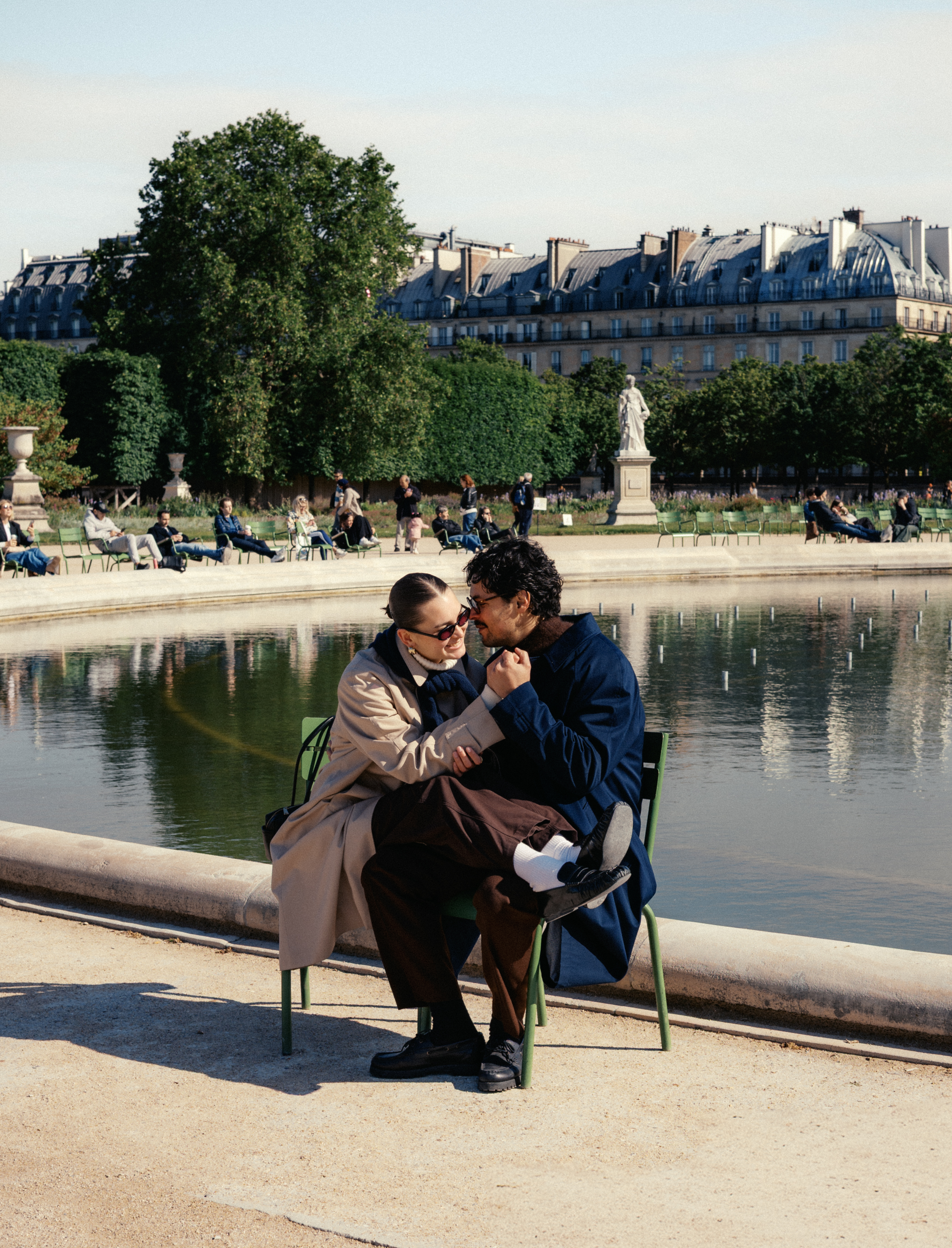 Couple sitting on a green bench by a reflecting pool with city buildings in the background.