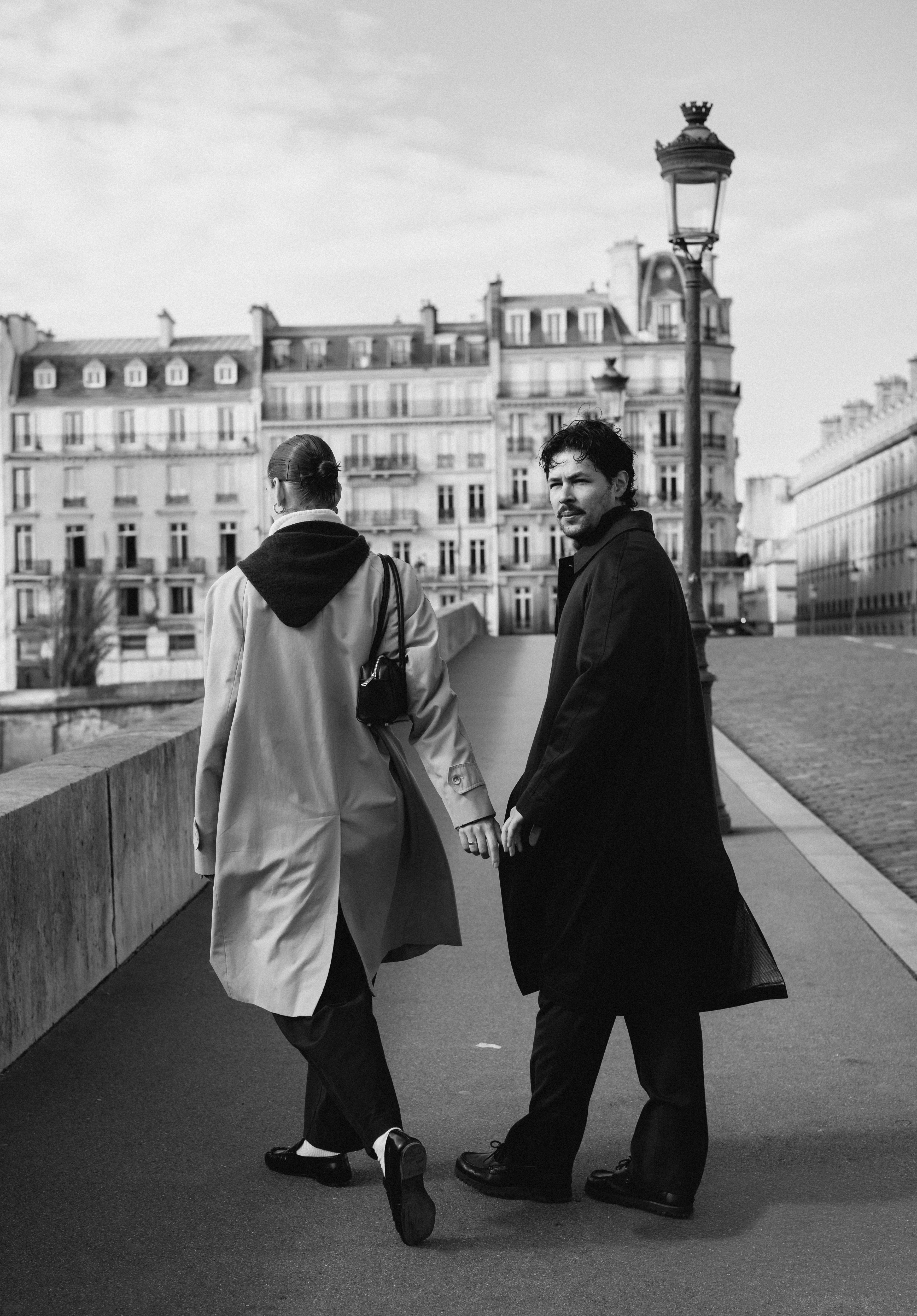 Two people walk hand in hand on a city sidewalk with Parisian buildings in the background (black and white).