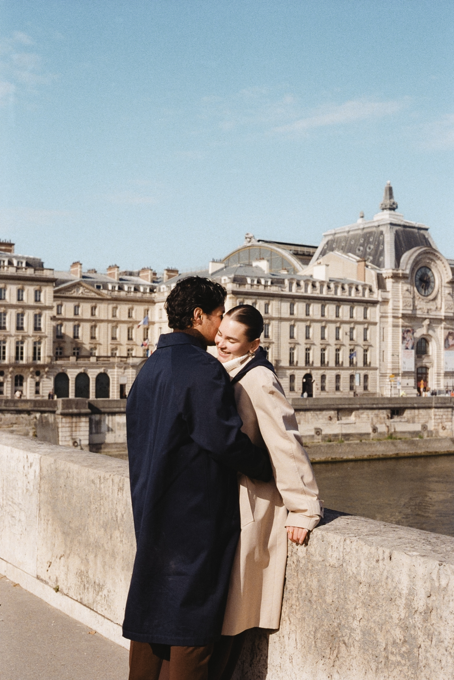 Couple embracing on a riverside promenade with historic buildings in the background.