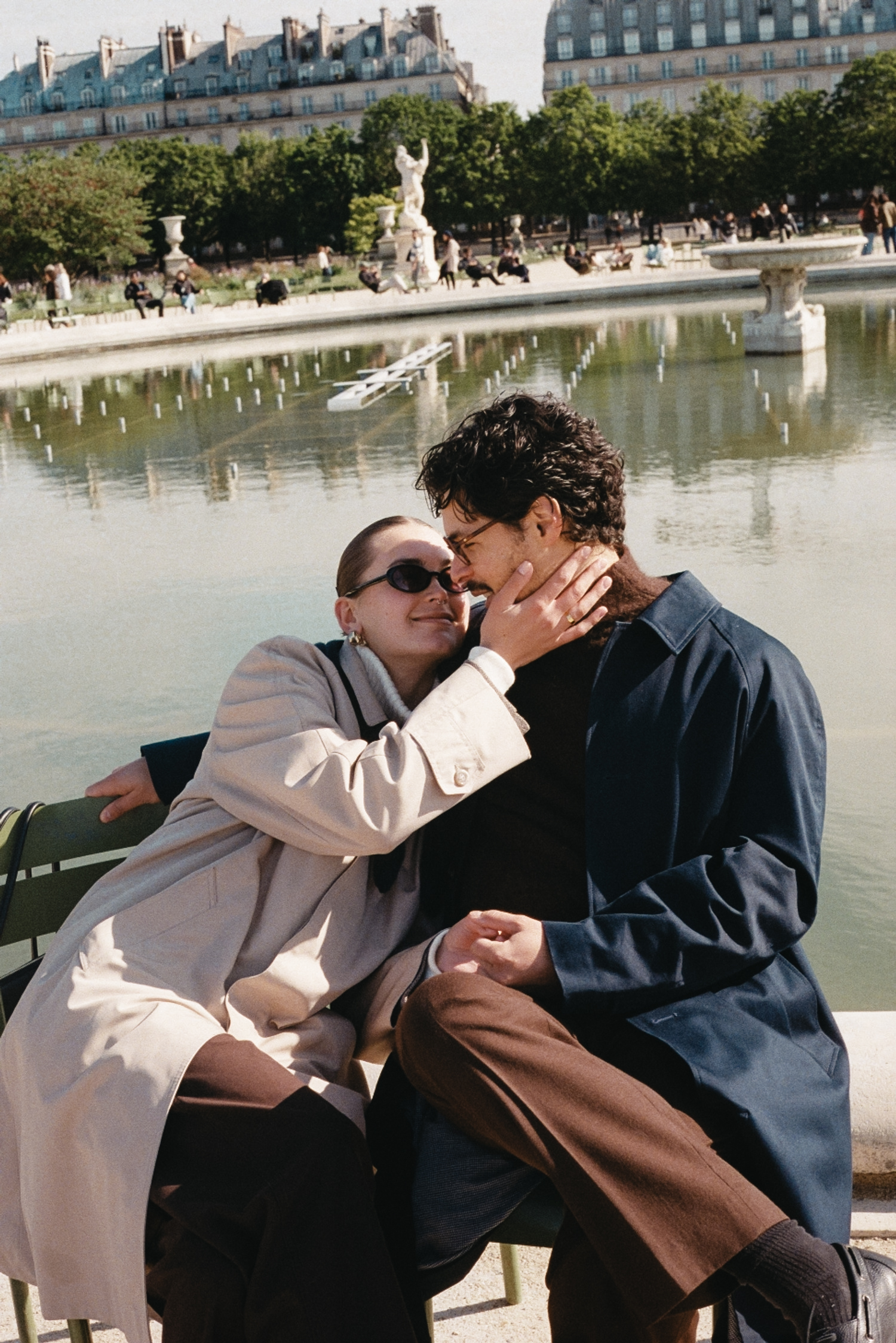 Couple sitting on bench by a fountain, woman wearing sunglasses and beige coat, man in dark jacket.