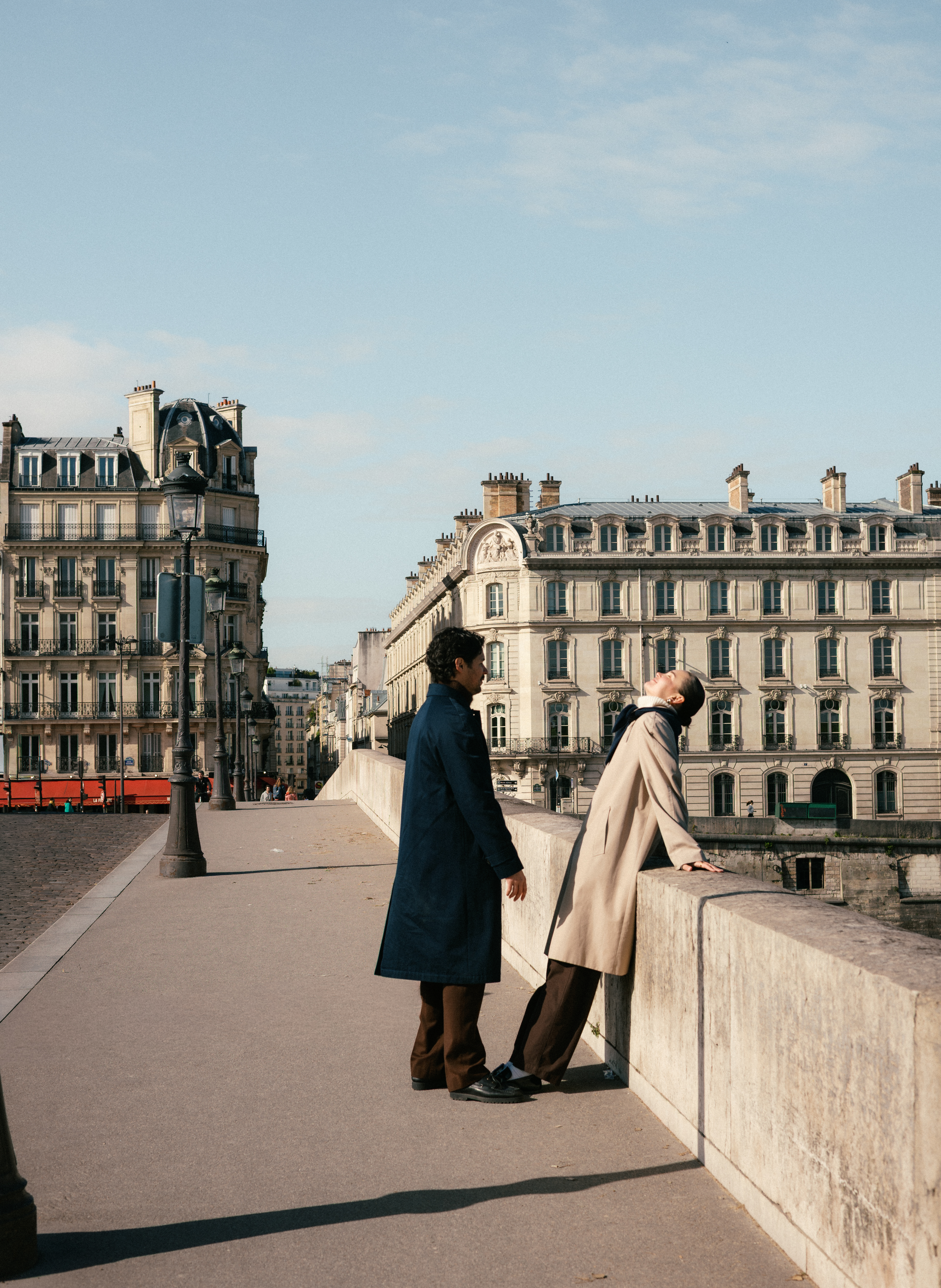 Two people stand on a riverfront promenade in Paris, facing opposite directions.
