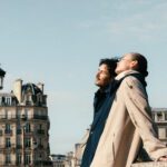 Couple in coats on a rooftop terrace with Parisian buildings and lampposts in clear sky