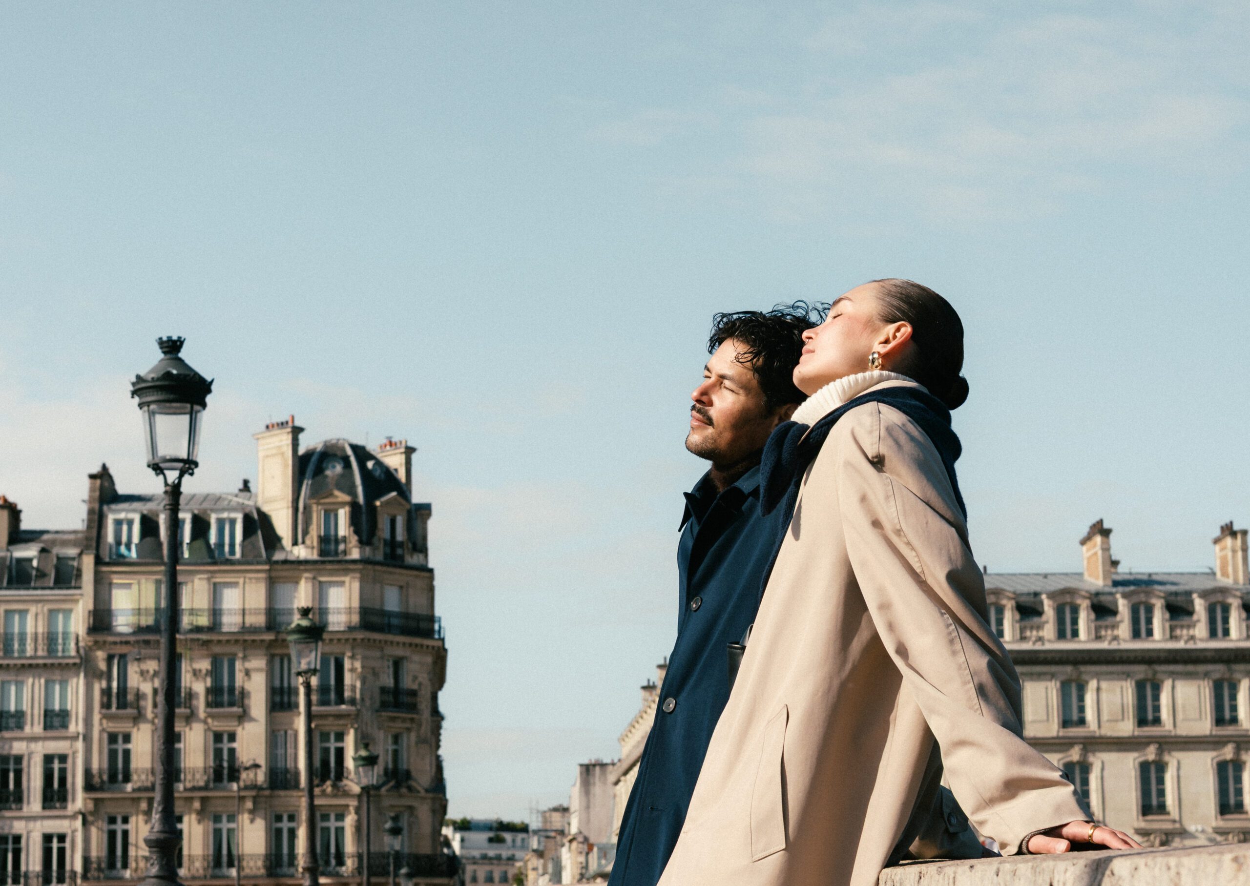 Couple in coats on a rooftop terrace with Parisian buildings and lampposts in clear sky