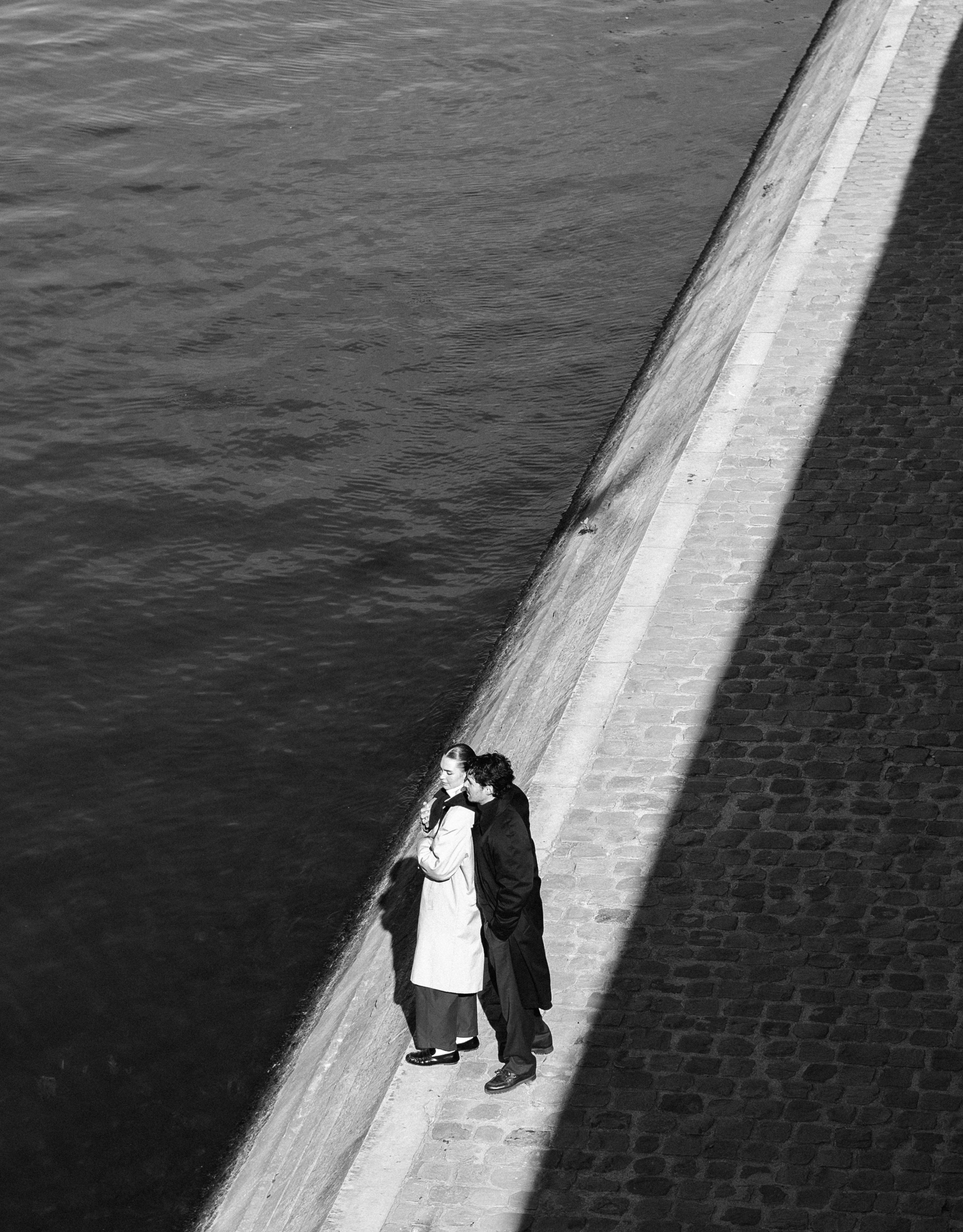 Two people stand on a stone walkway beside a body of water, with a diagonal shadow across the scene.