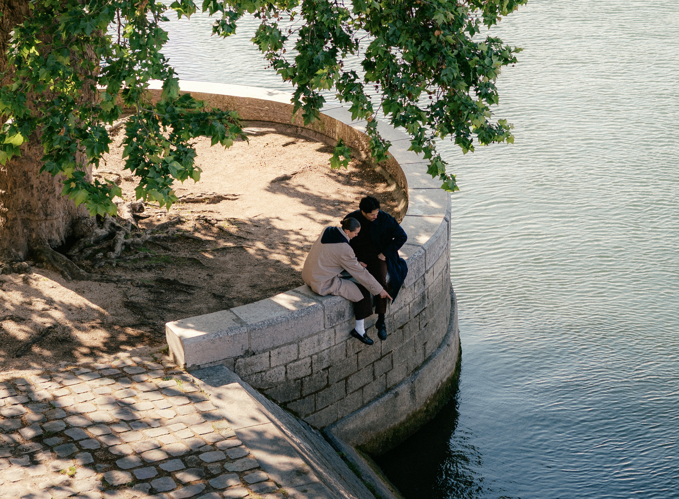 Two people sitting on a curved stone ledge by a calm river, shaded by overhanging tree branches.