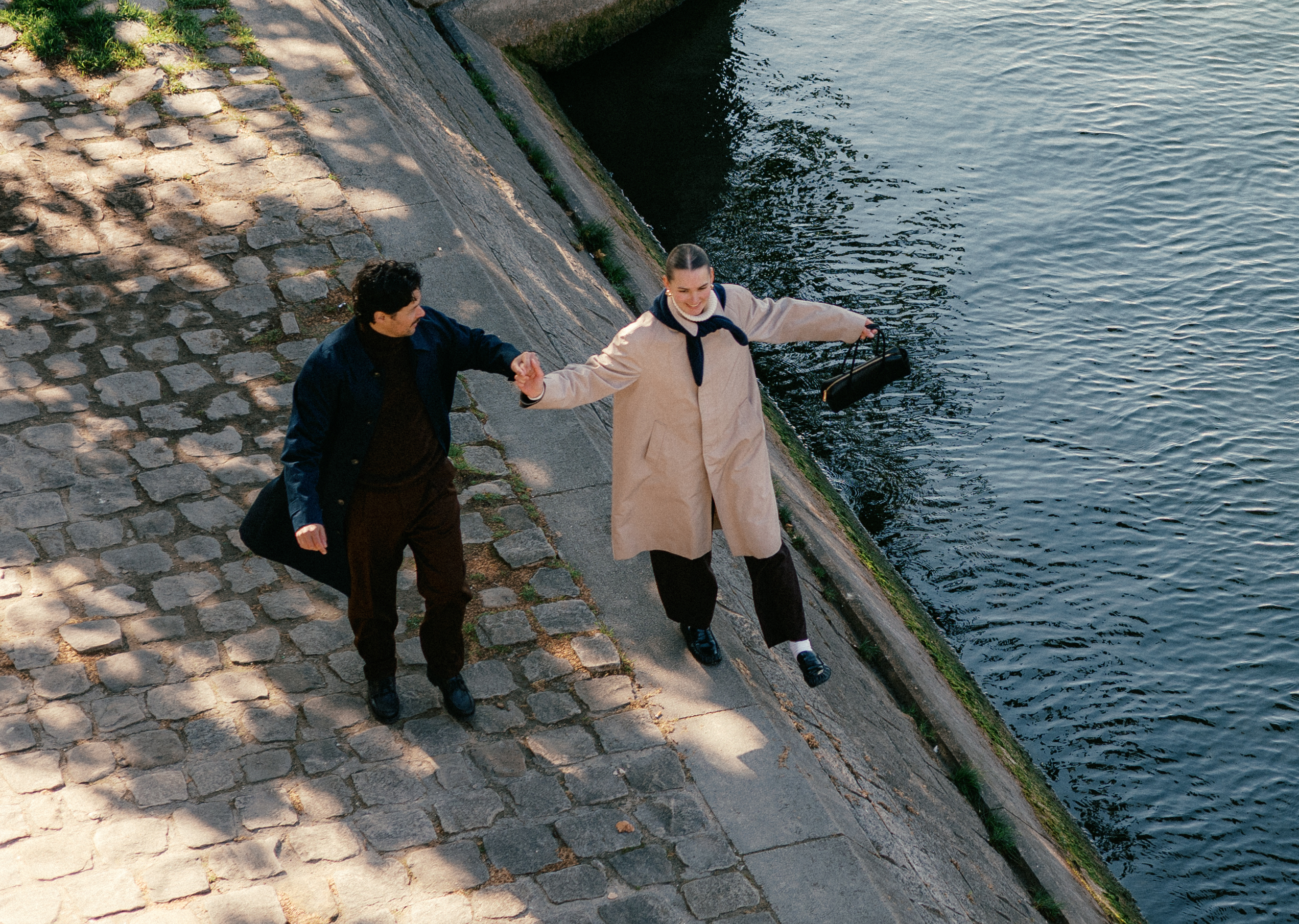 Two people walk along a sloped stone path by a calm body of water, holding hands.