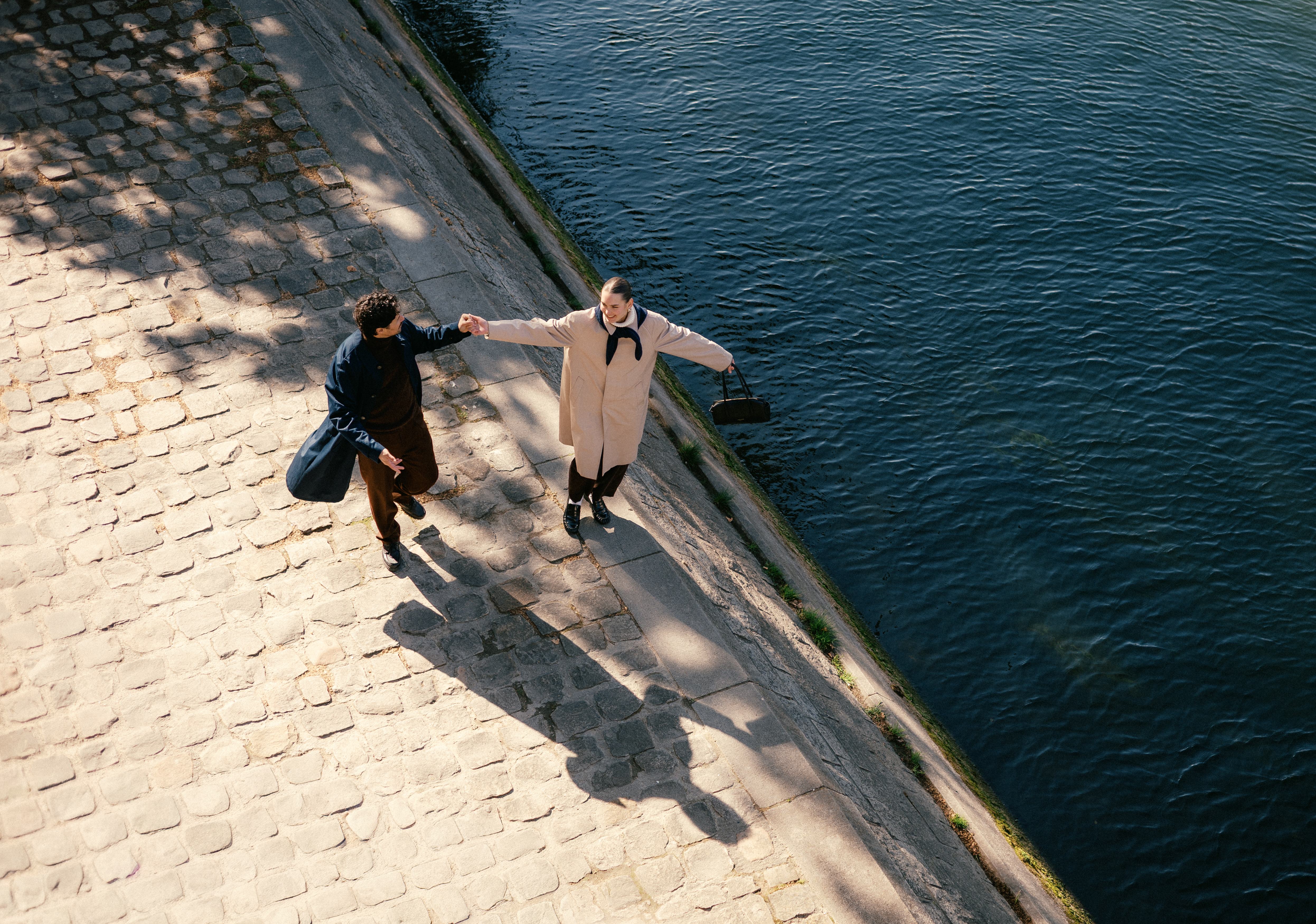 Two people walking along a stone embankment by water, holding hands; shadows on path.