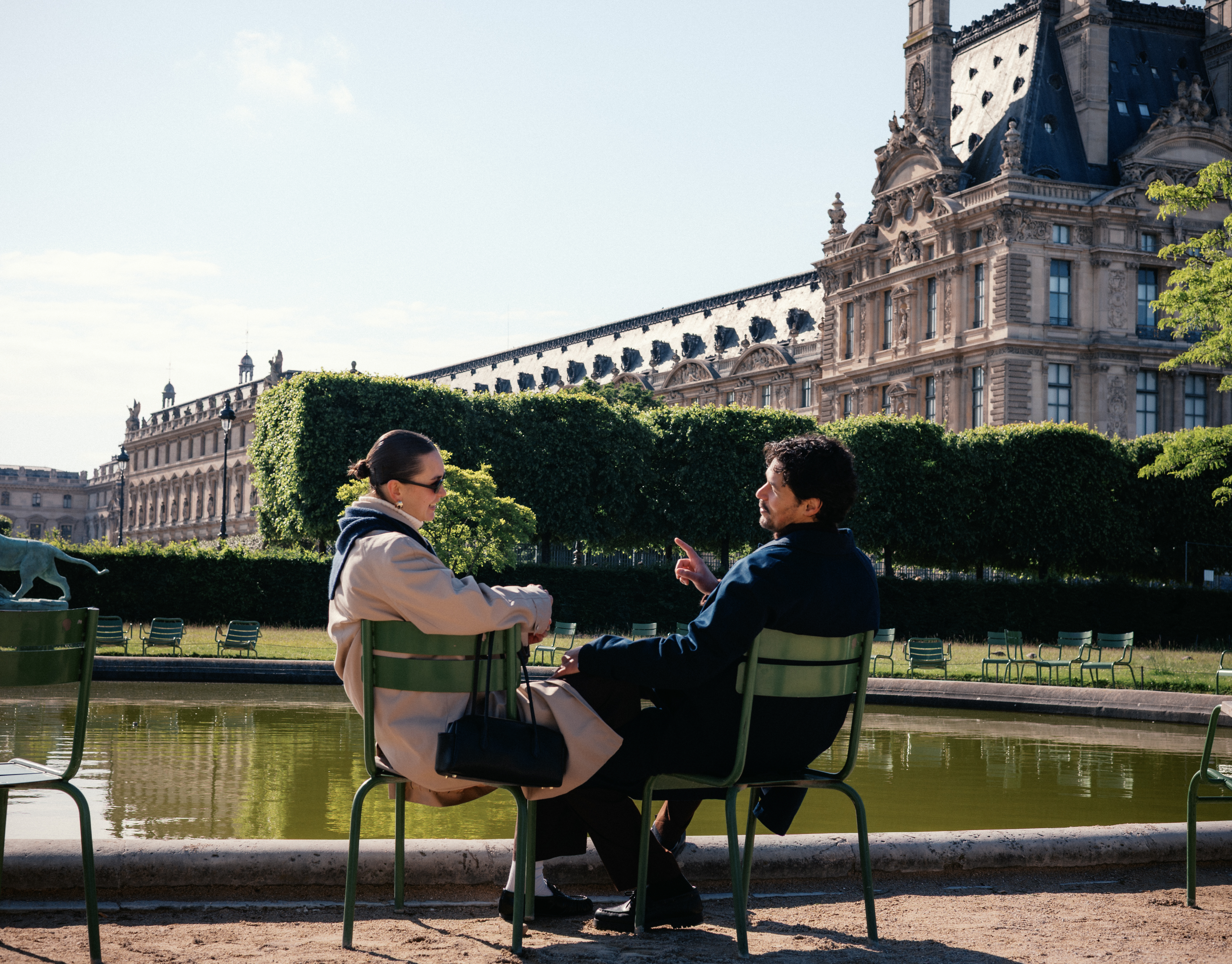Two people sit and converse on green chairs beside a reflective pool with a historic ornate building in the background.