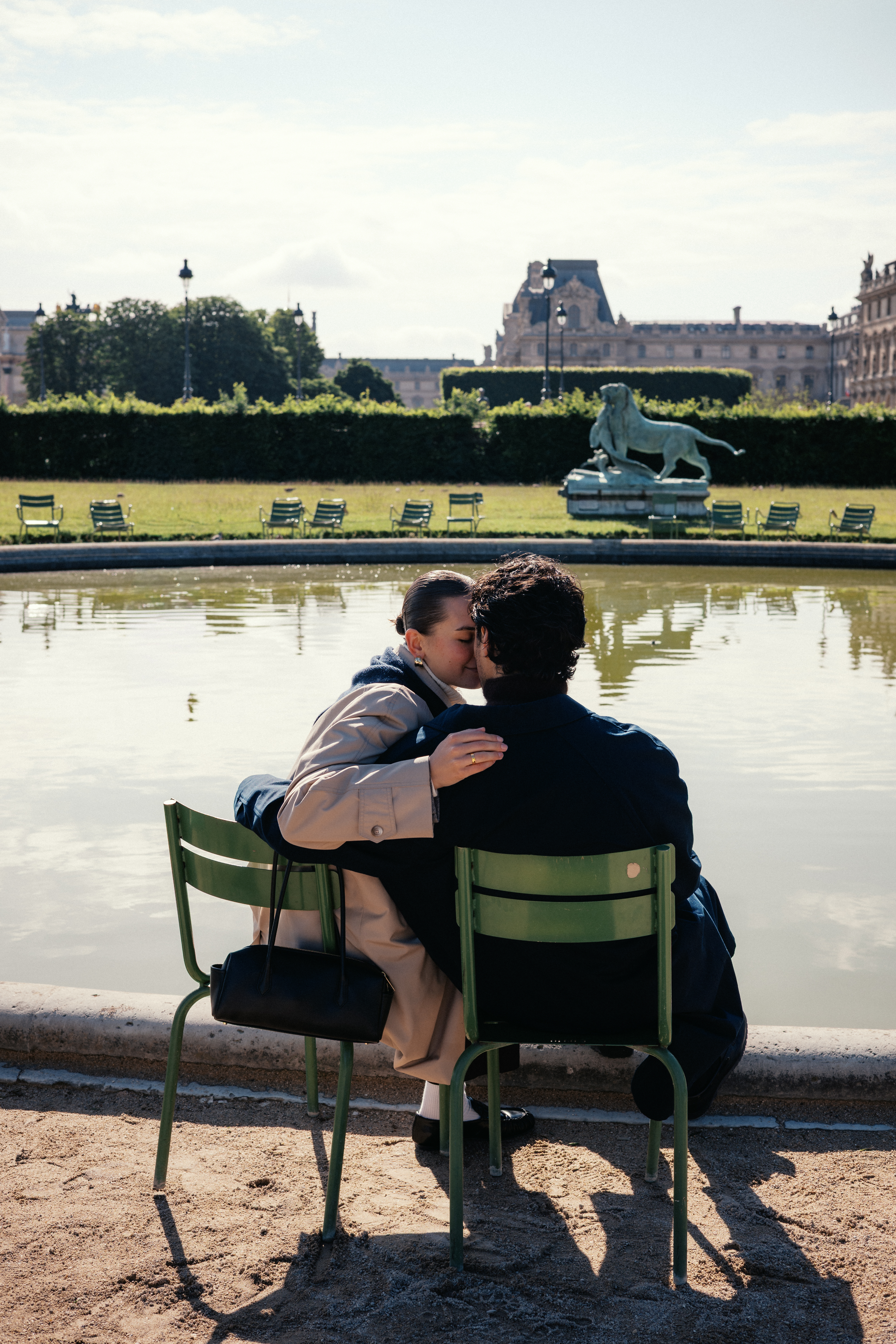 Couple kissing on green chairs by a fountain with statues and historic buildings in background