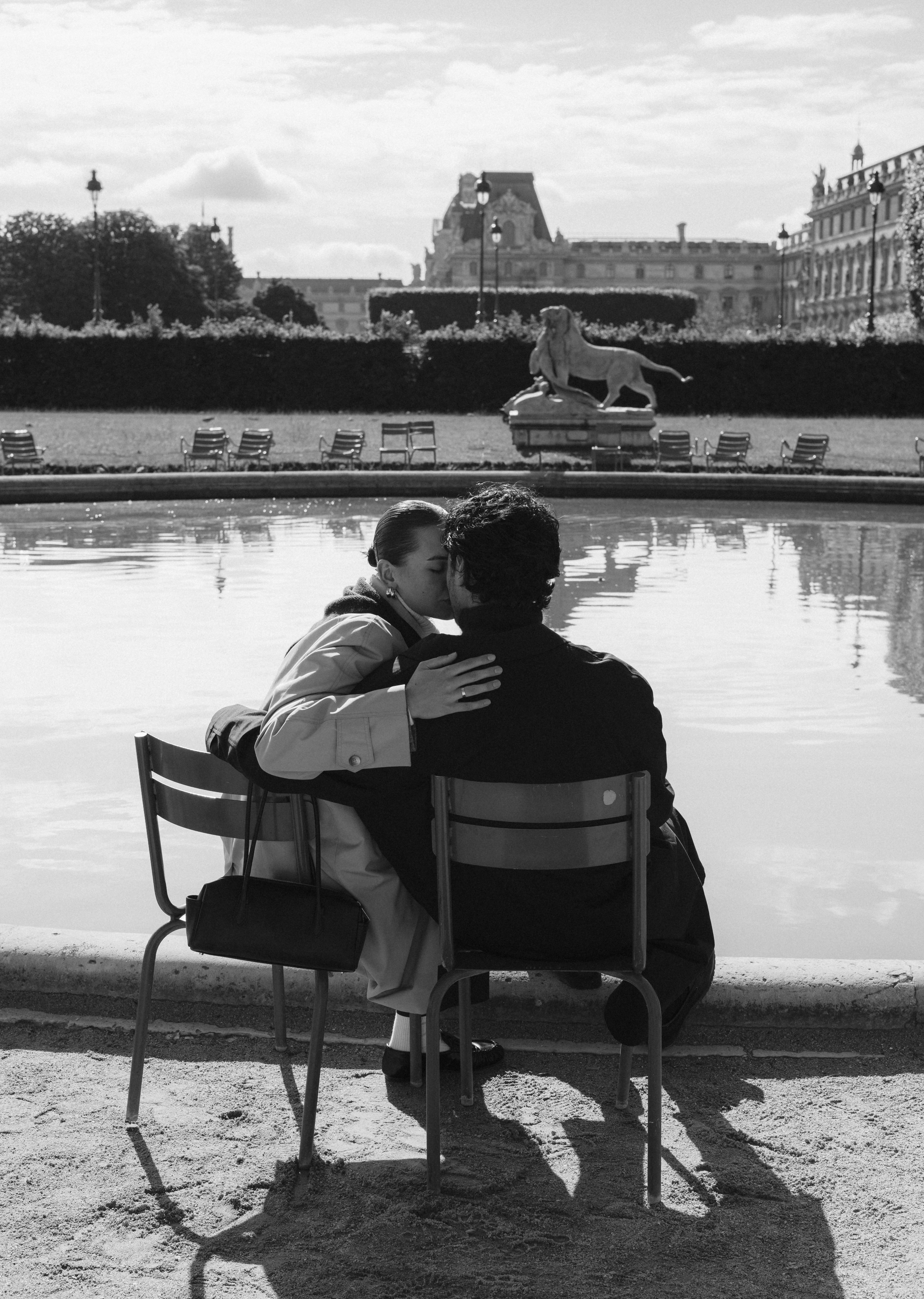 Couple kissing on chairs by a reflective pool with statue and historic buildings in background.