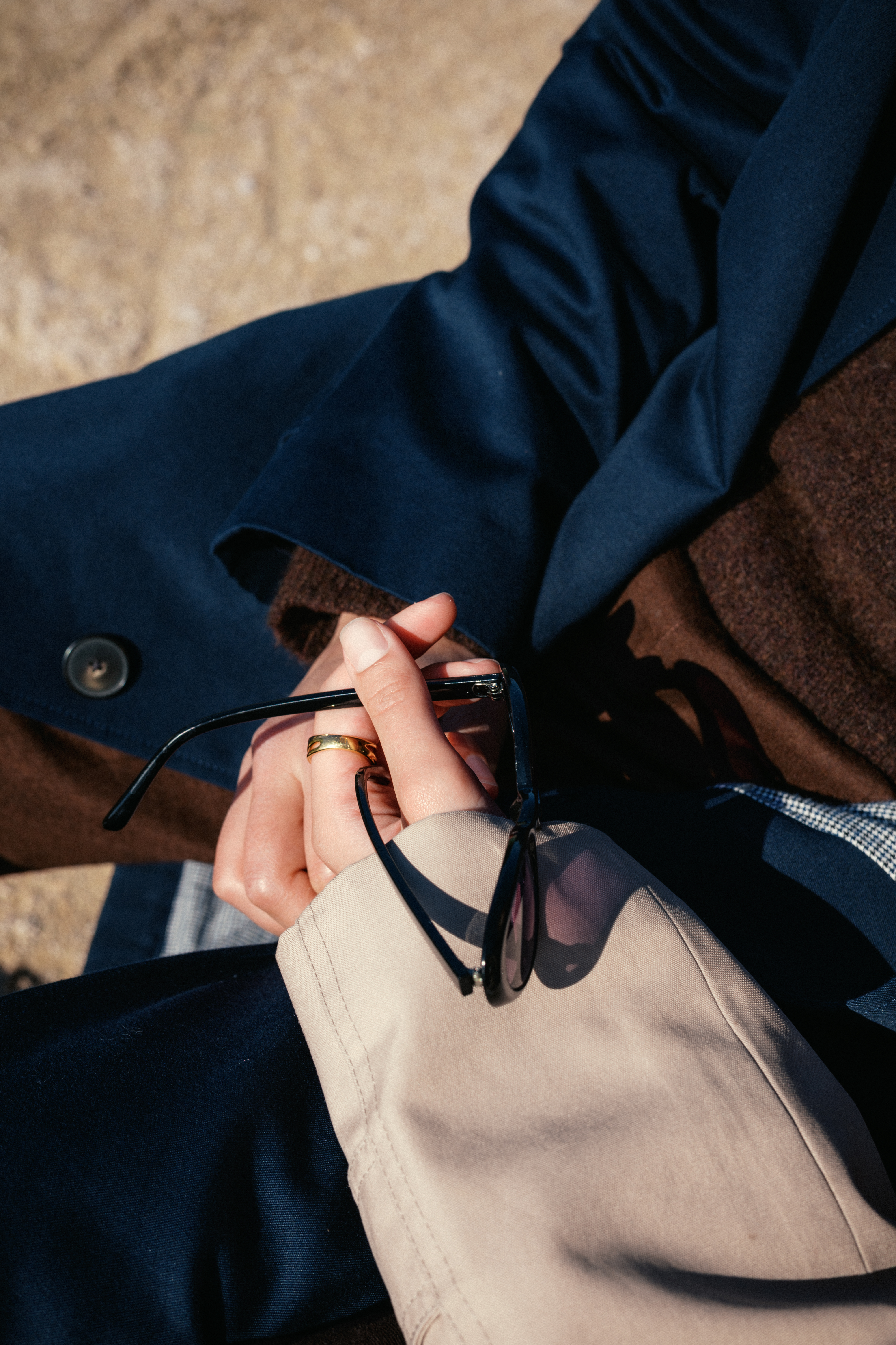 Close-up of hands holding black-framed glasses over a beige bag, navy coat, and brown garment.