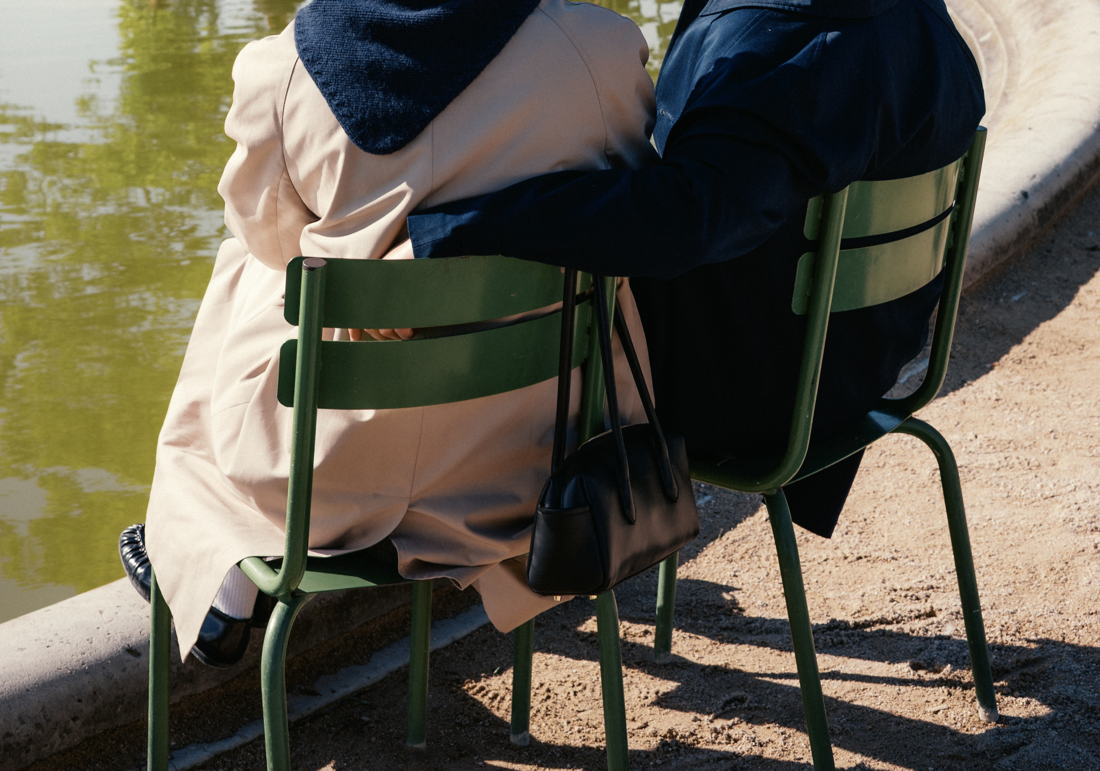 Two people sit on green chairs by a pond, arms around each other, bags beside.