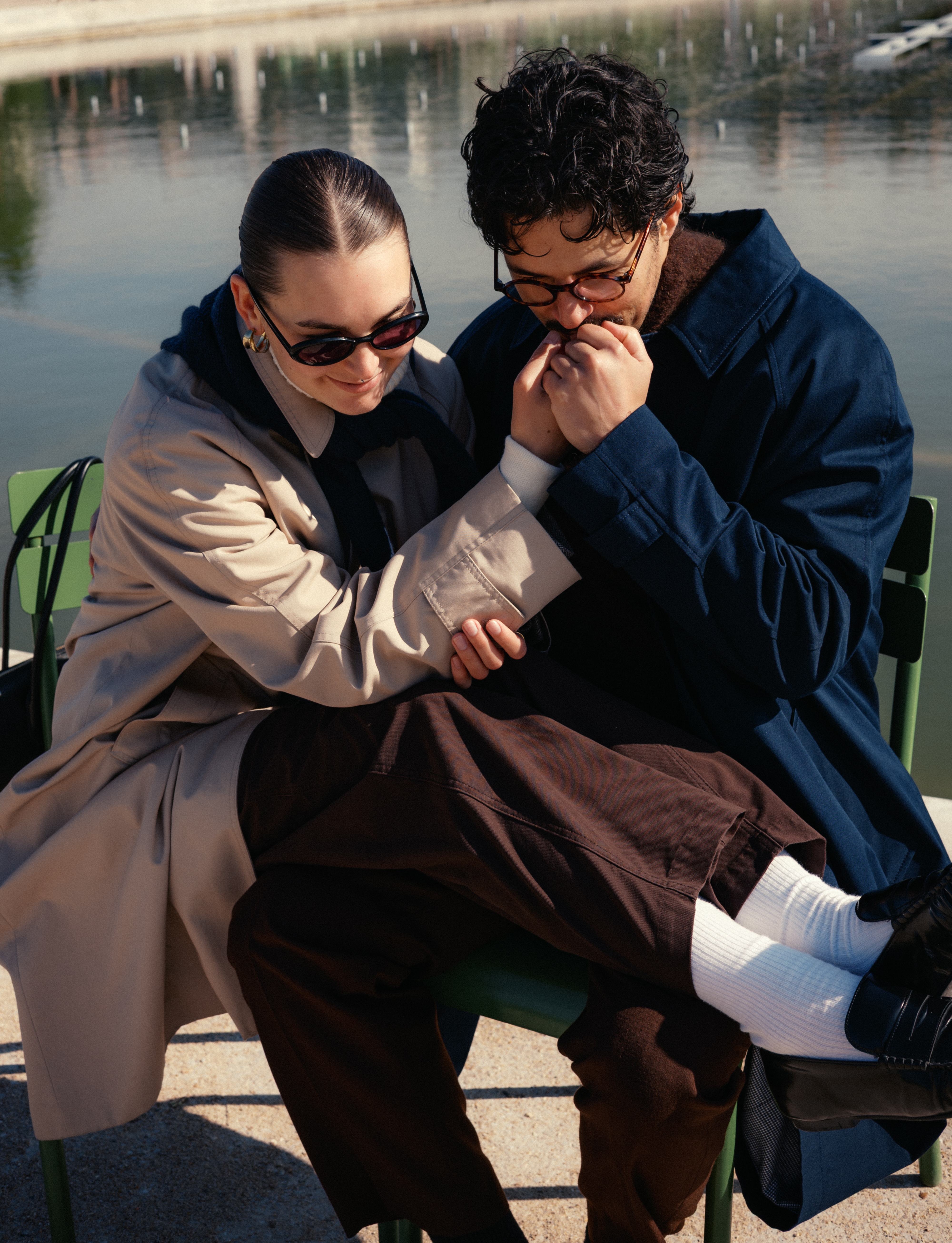 Two people sit closely on a green bench by a body of water, wearing coats and sunglasses.