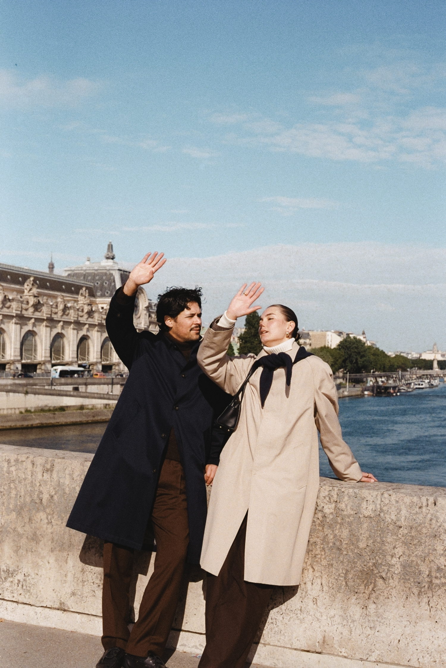 Two people in long coats raise hands by a stone railing along a river, Musée d'Orsay building in background.