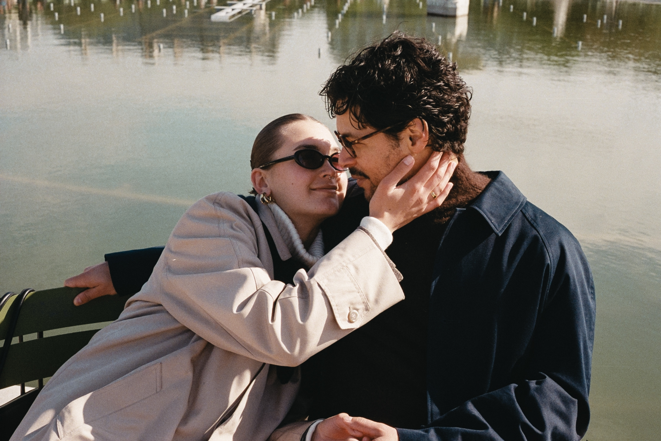 Couple embracing on a boat by water; woman wears beige coat, man in dark jacket, sunglasses.
