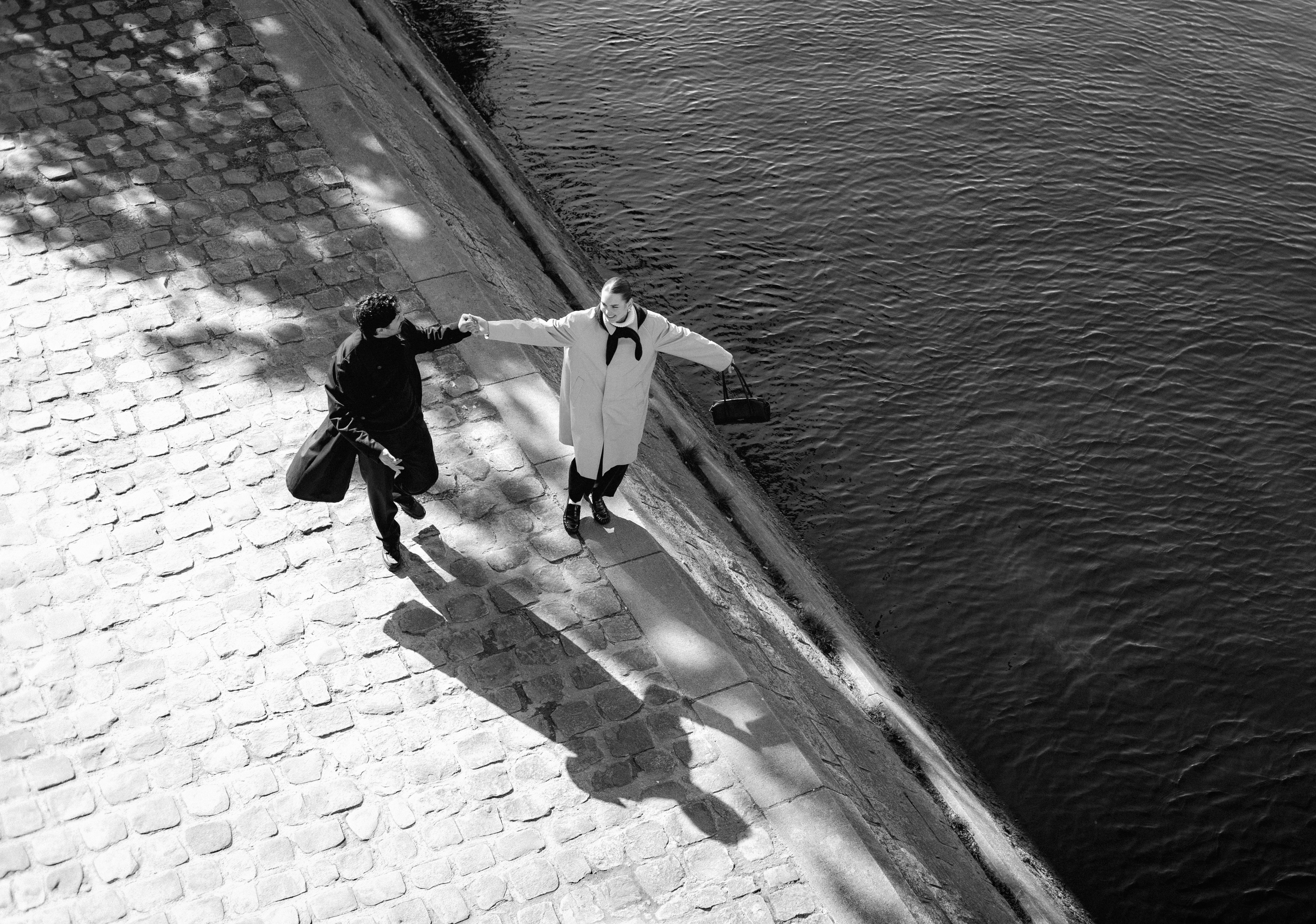 Two people on a stone walkway by water, holding hands; dappled shadows on cobbles.
