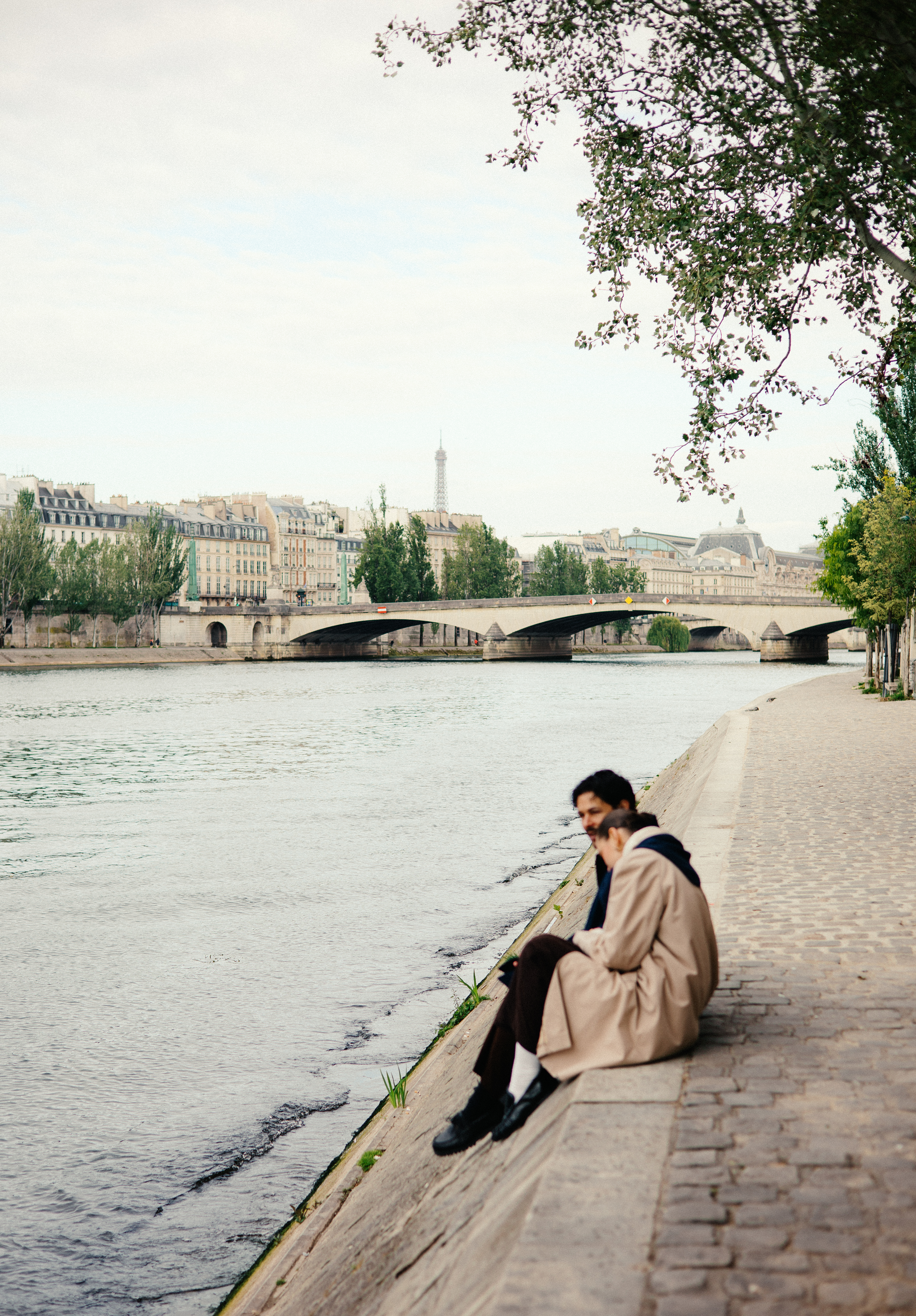 Riverside promenade with buildings, bridge, and people sitting by the water along a stone embankment.