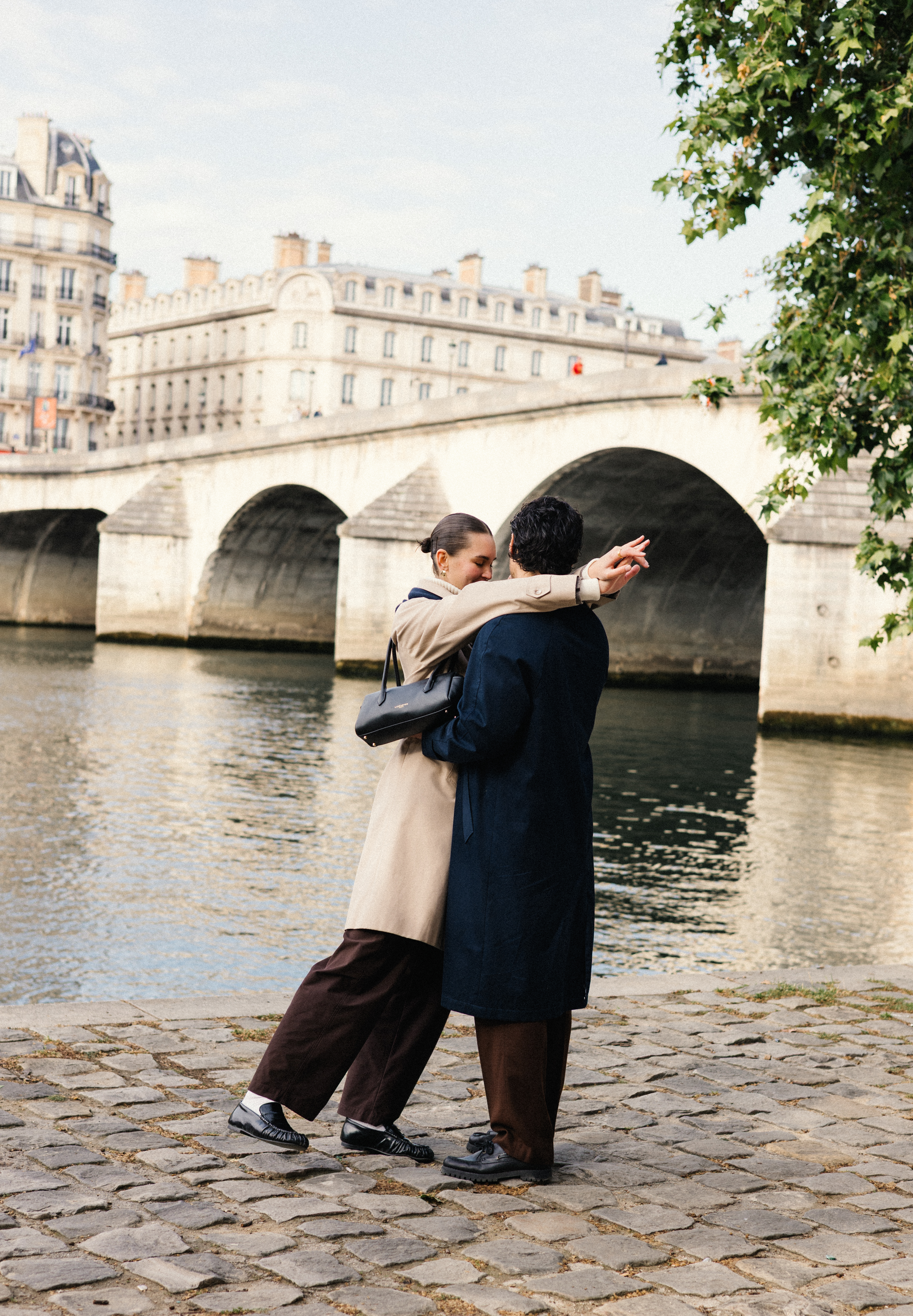 Couple embracing by a river with stone bridge and Parisian buildings in background.