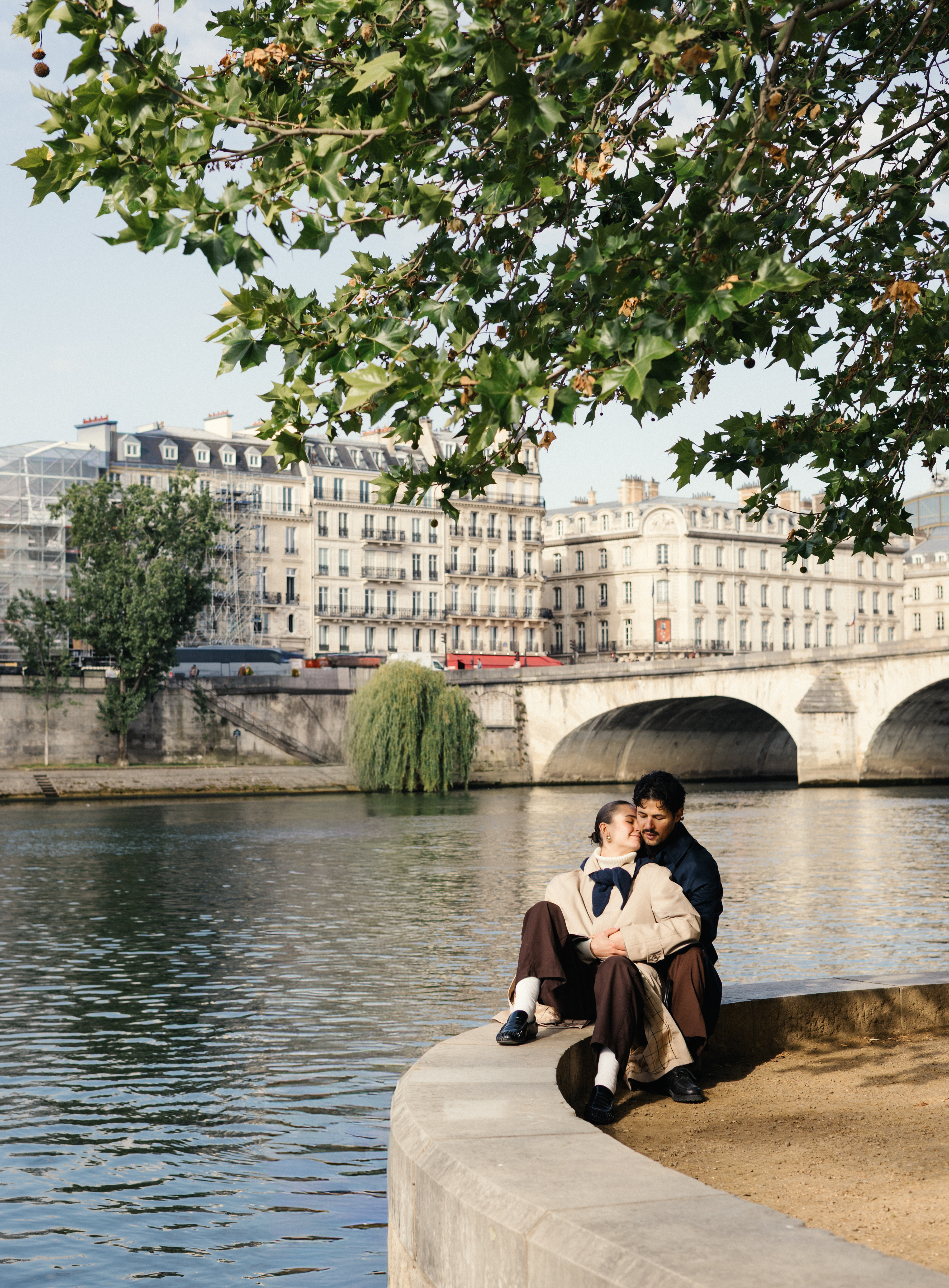 Couple sitting on riverbank ledge with city buildings and bridge behind them.