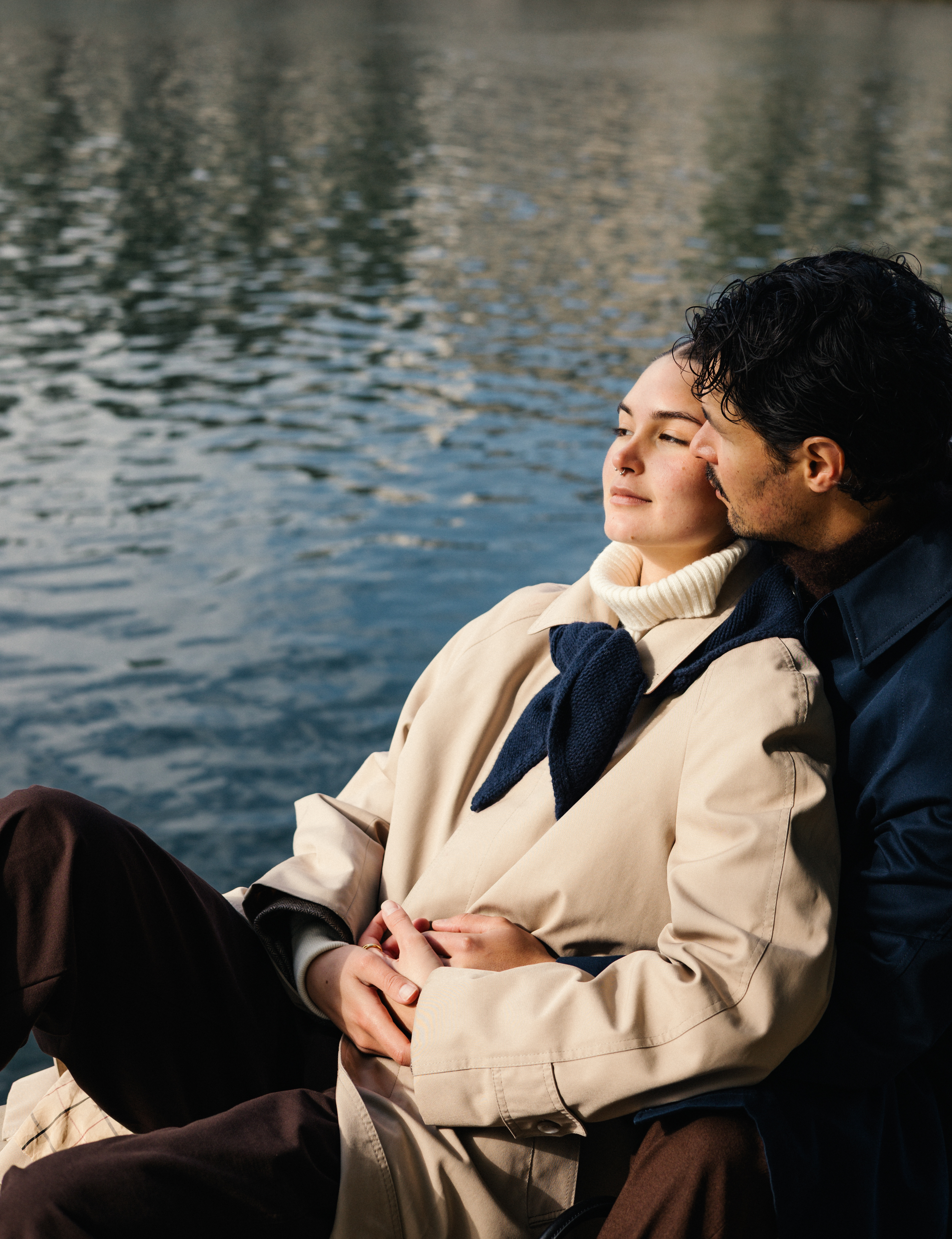 Two people sit by a lake; a woman rests with eyes closed while a man leans beside her.