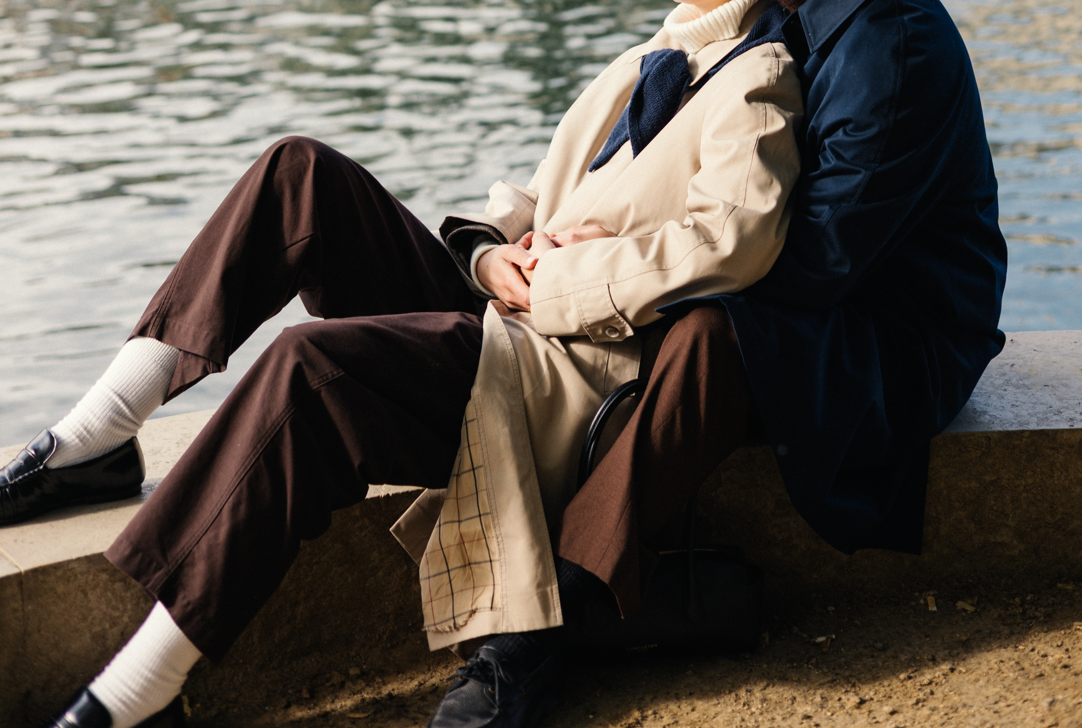 Two people sit on a stone ledge by water, wearing beige coat and navy jacket.
