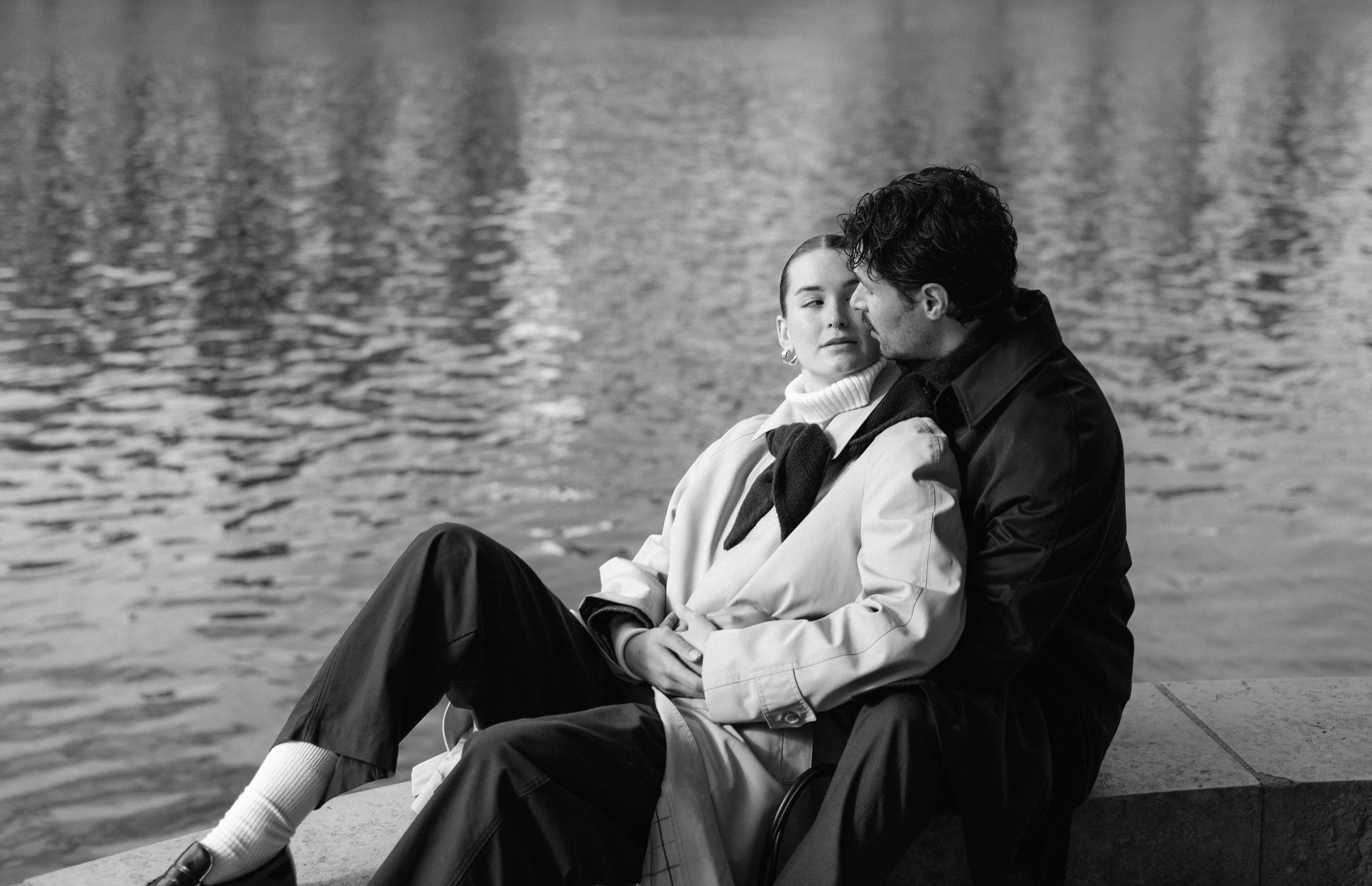 Couple sitting by water; man behind woman, both looking at each other in black-and-white