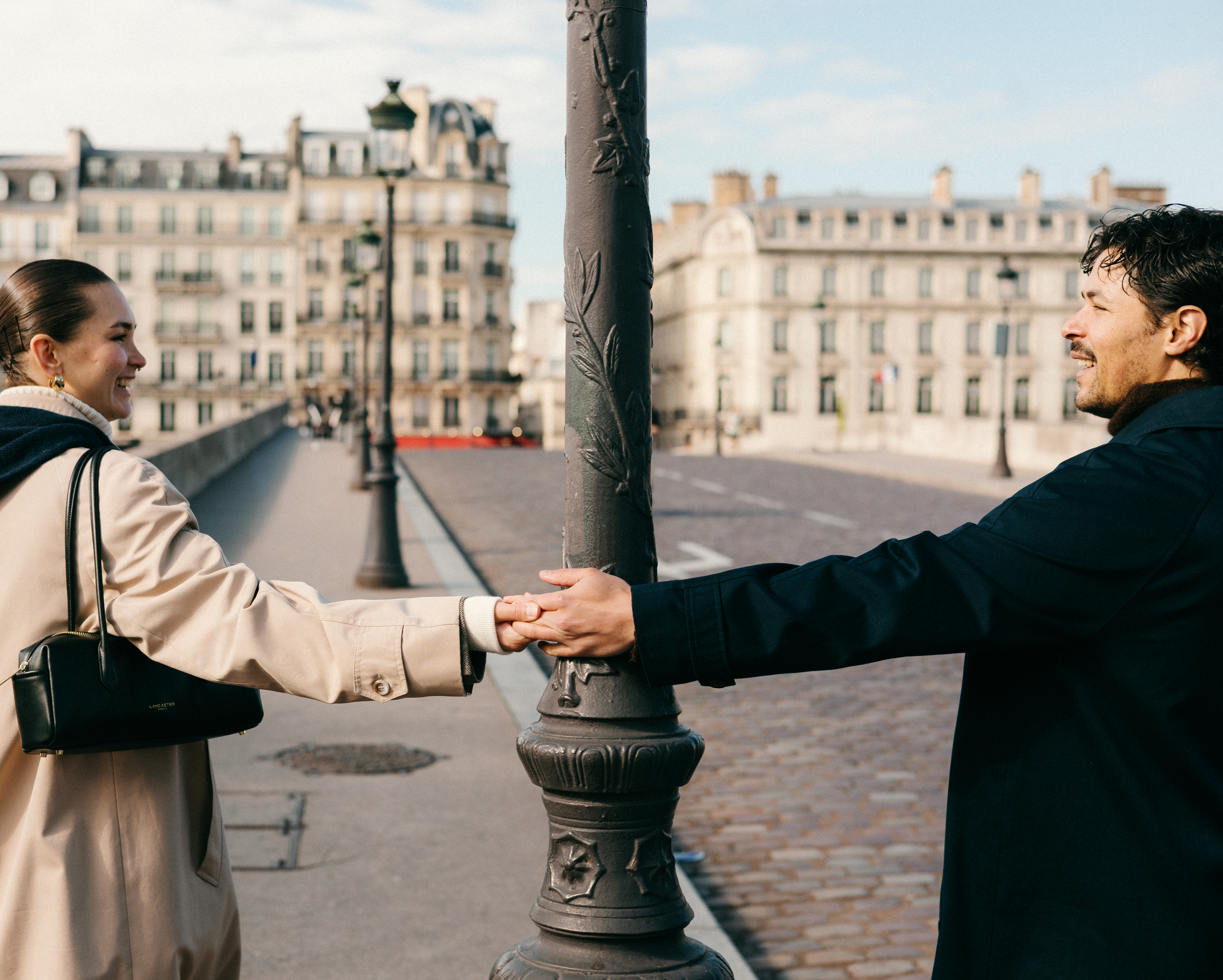 Two people hold hands around a lamppost on a city street with historic buildings in background.