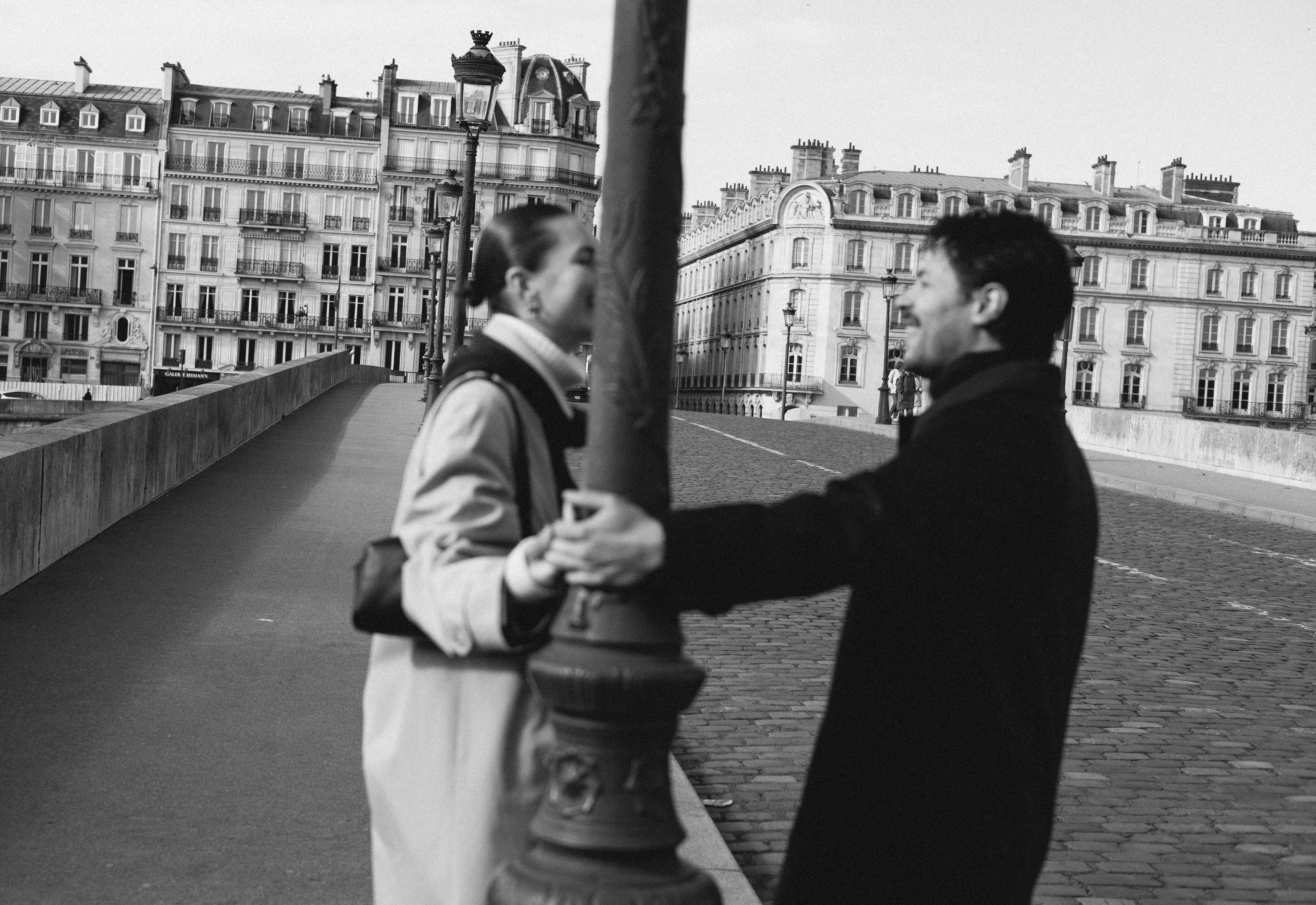 Two people embrace and kiss on a cobbled street with Parisian buildings in the background (black and white).