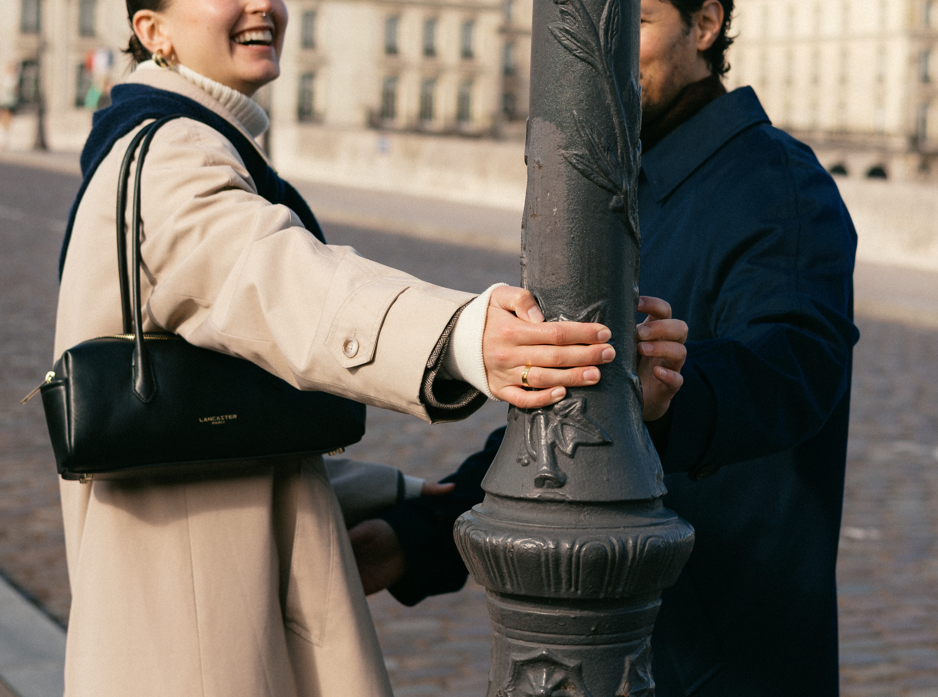Two people stand on a street and hold a lamppost between them, smiling and leaning.