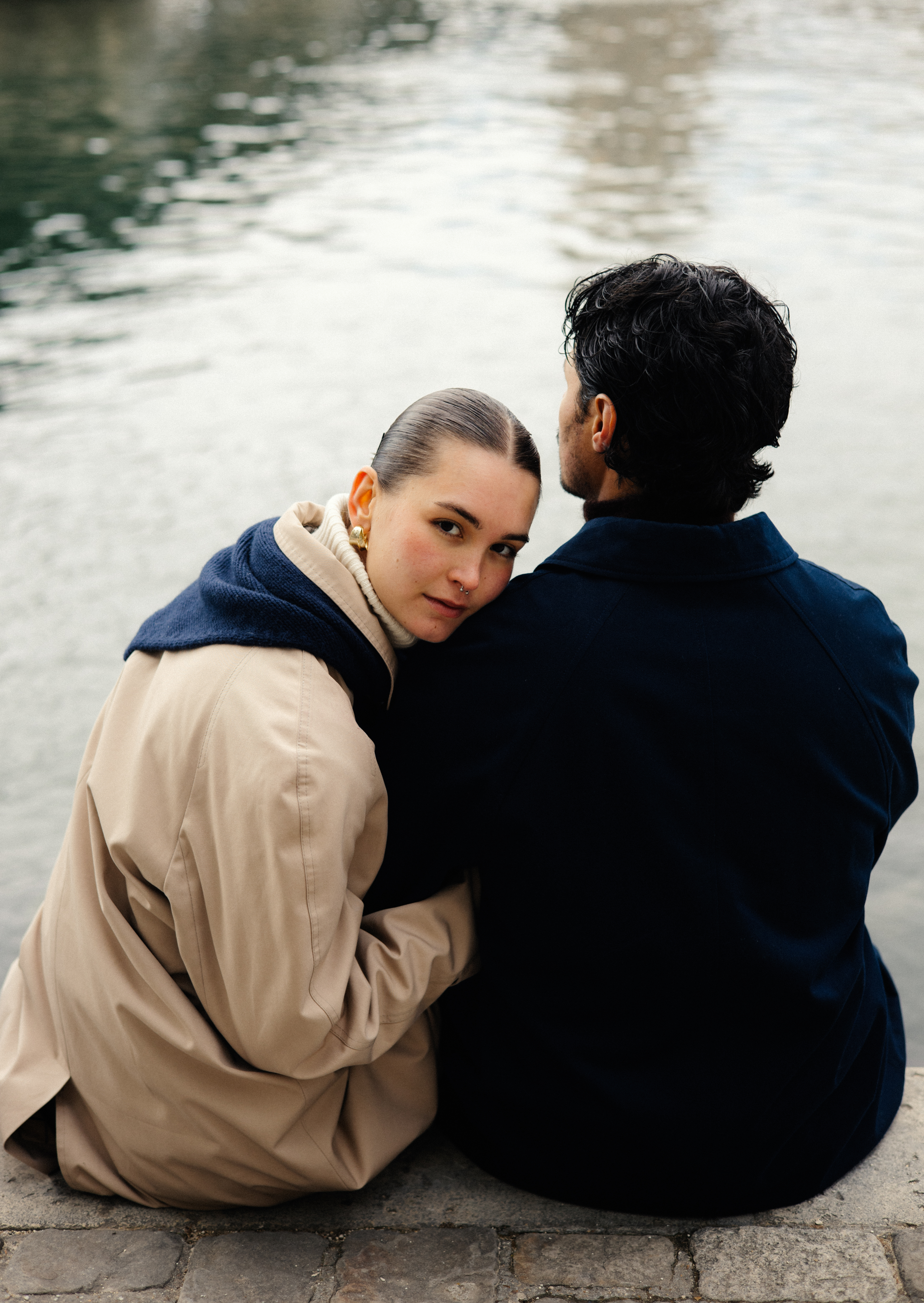 Two people sit together by a body of water, one resting head on the other's shoulder.