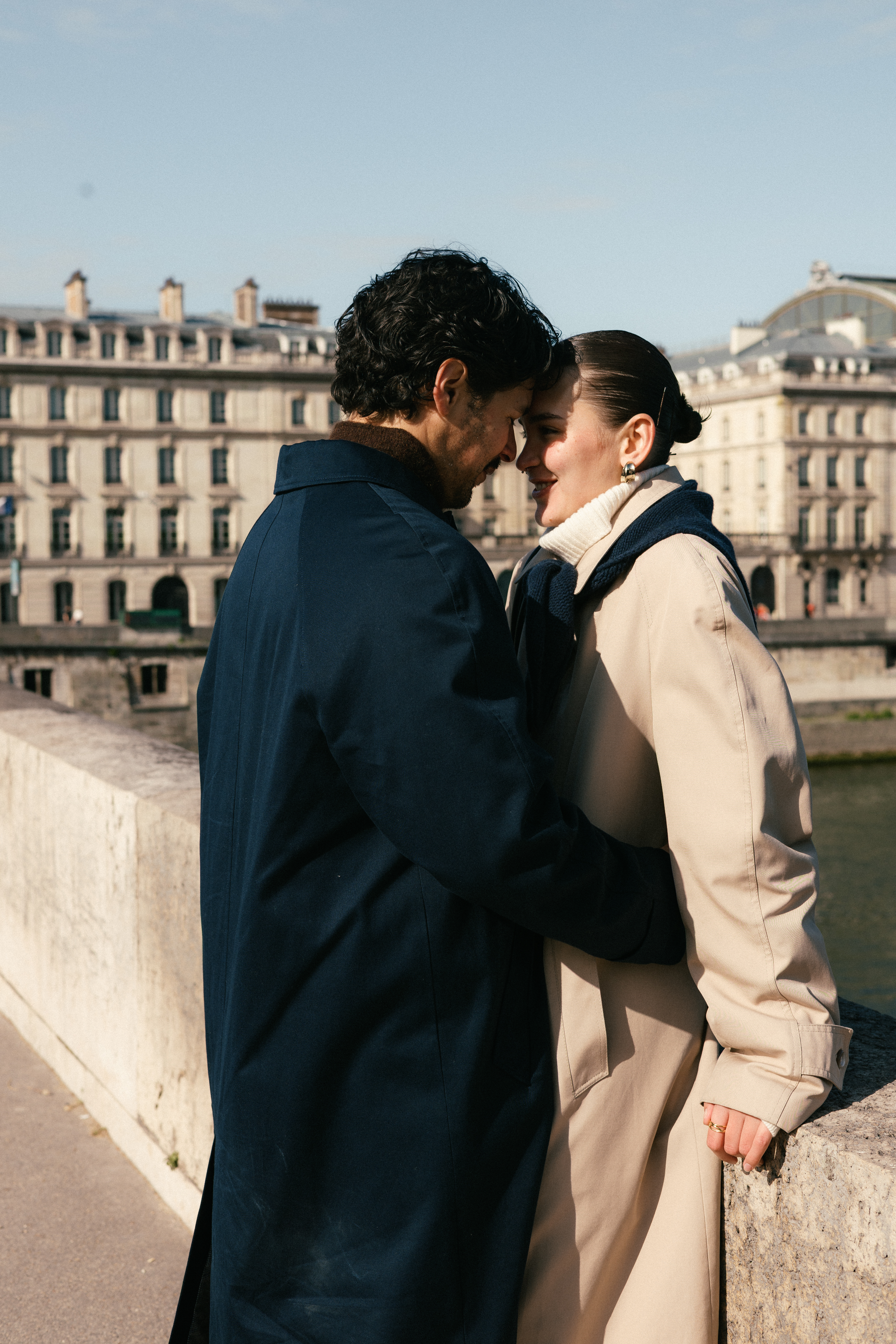 Two people in coats stand close, foreheads touching near a riverside urban backdrop.