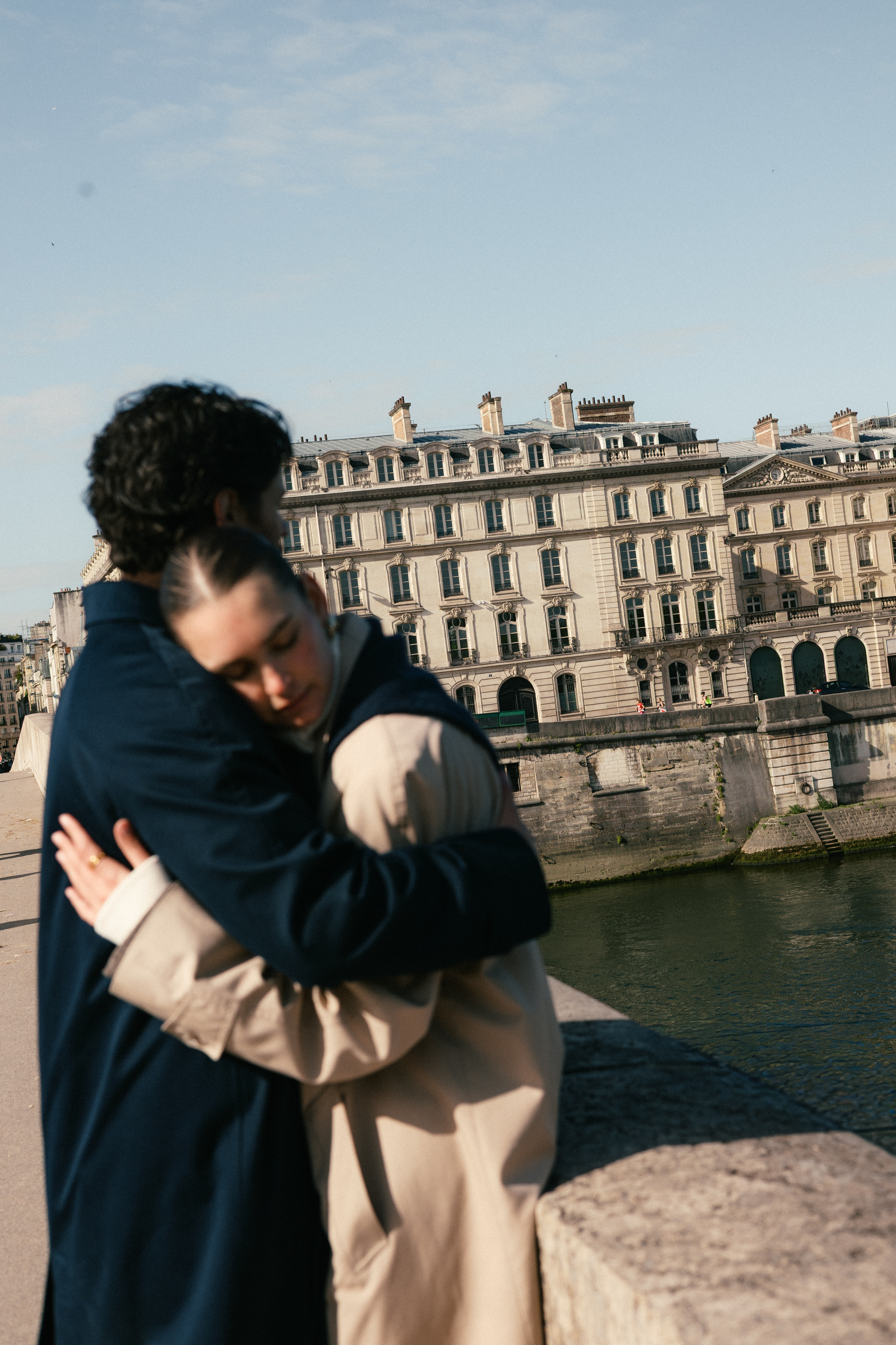 Two people embrace near a river, with ornate Parisian-style buildings in the background.