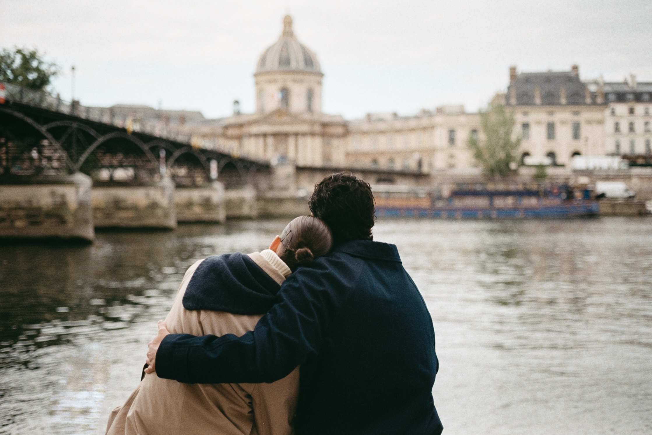 Two people embrace by a river with a cityscape featuring a domed building and bridge in the background.