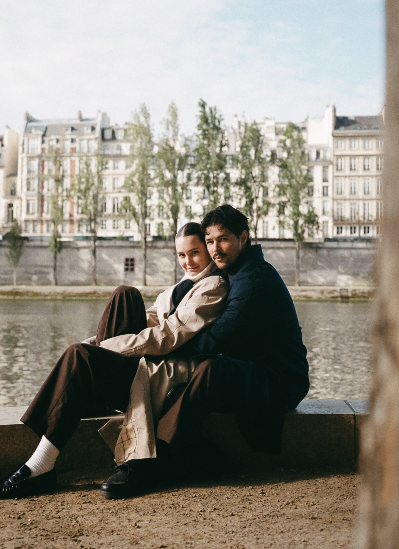 Two people sit closely by a river, Parisian buildings in the background.