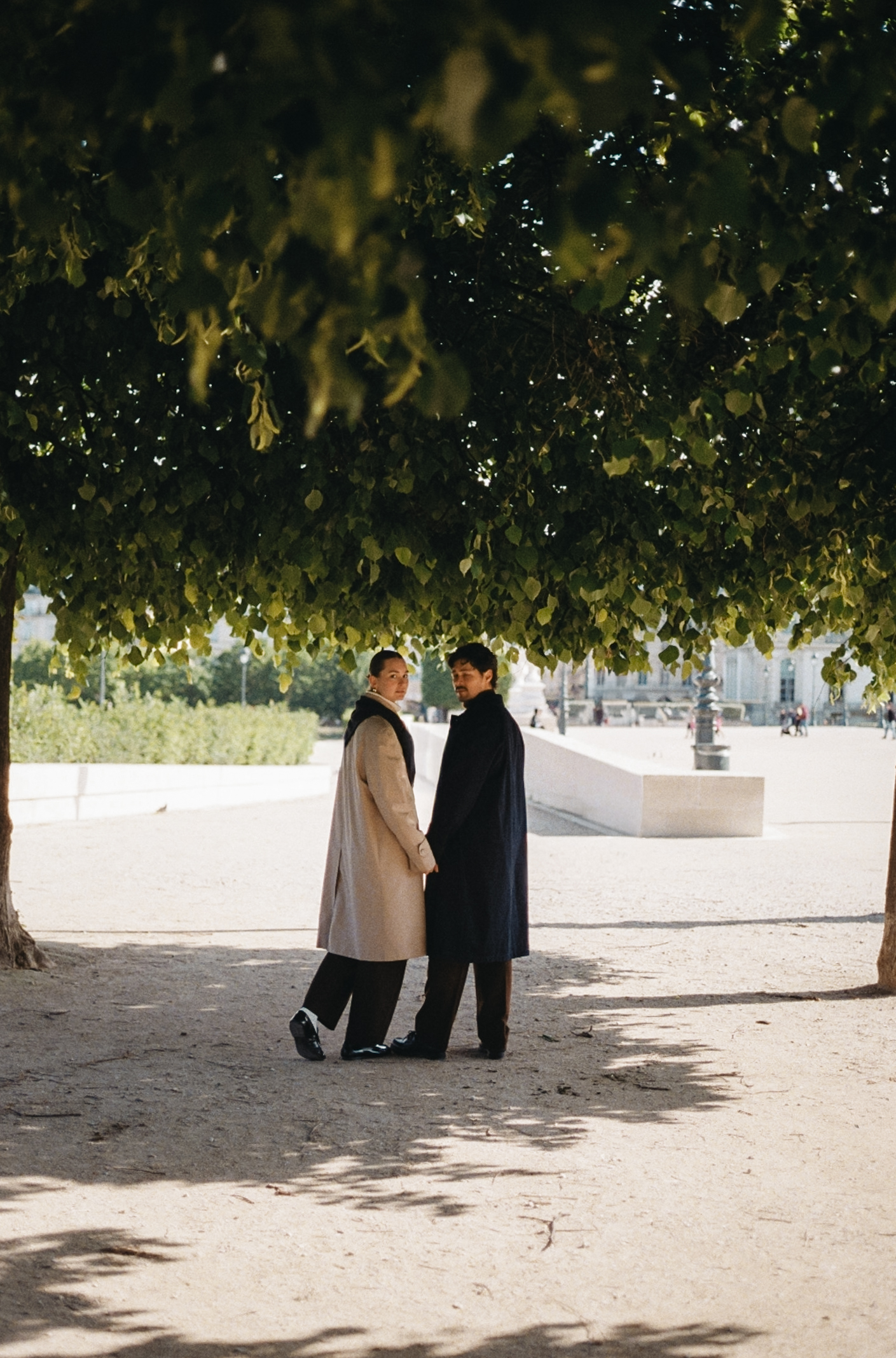 Two people stand under a leafy canopy, facing each other and holding hands in a sunny outdoor setting.