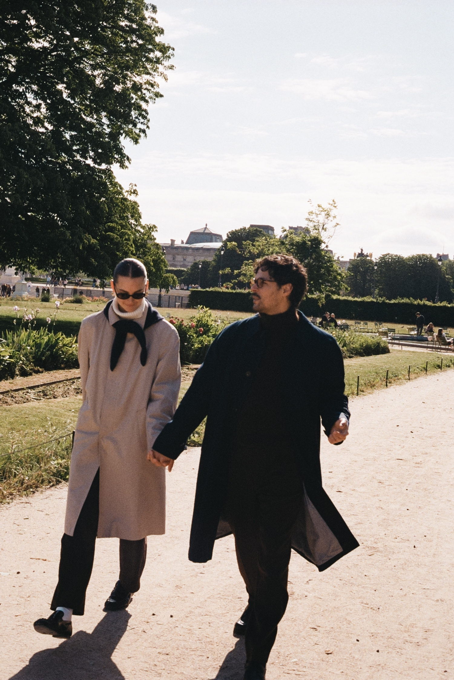 Two people walk hand in hand along a sunlit park path with trees and shrubs in the background.