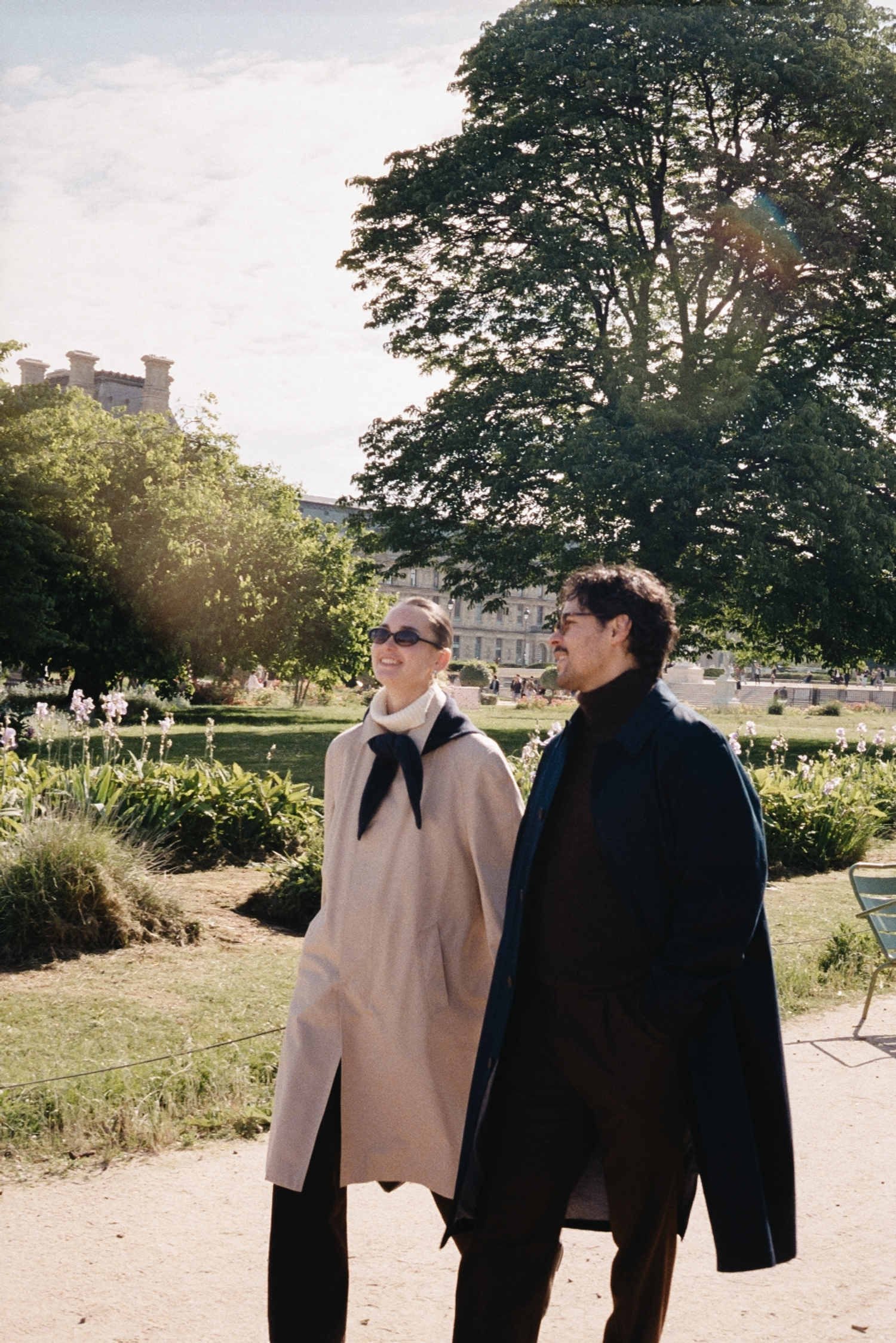 Two people walking outdoors in a park, smiling, wearing coats and sunglasses.