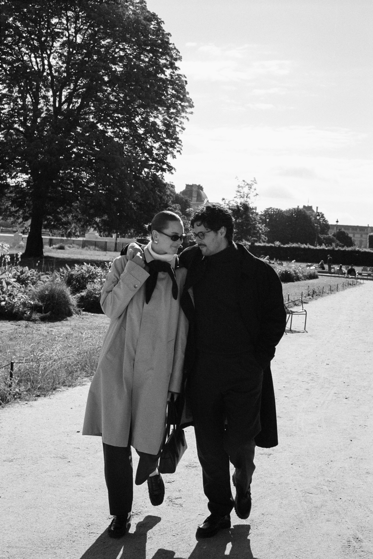 Two people walking arm in arm on a sunny day in a park, wearing coats.