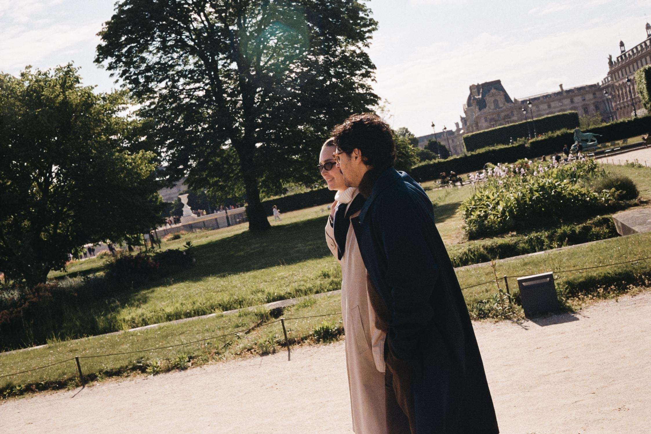 Couple walking in a park near the Louvre, trees, shrubs, and gravel paths.