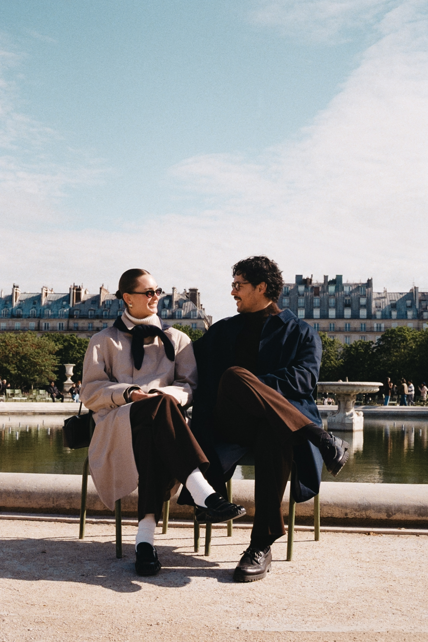 Two people sit on a bench by a fountain in a Parisian park, smiling at each other.